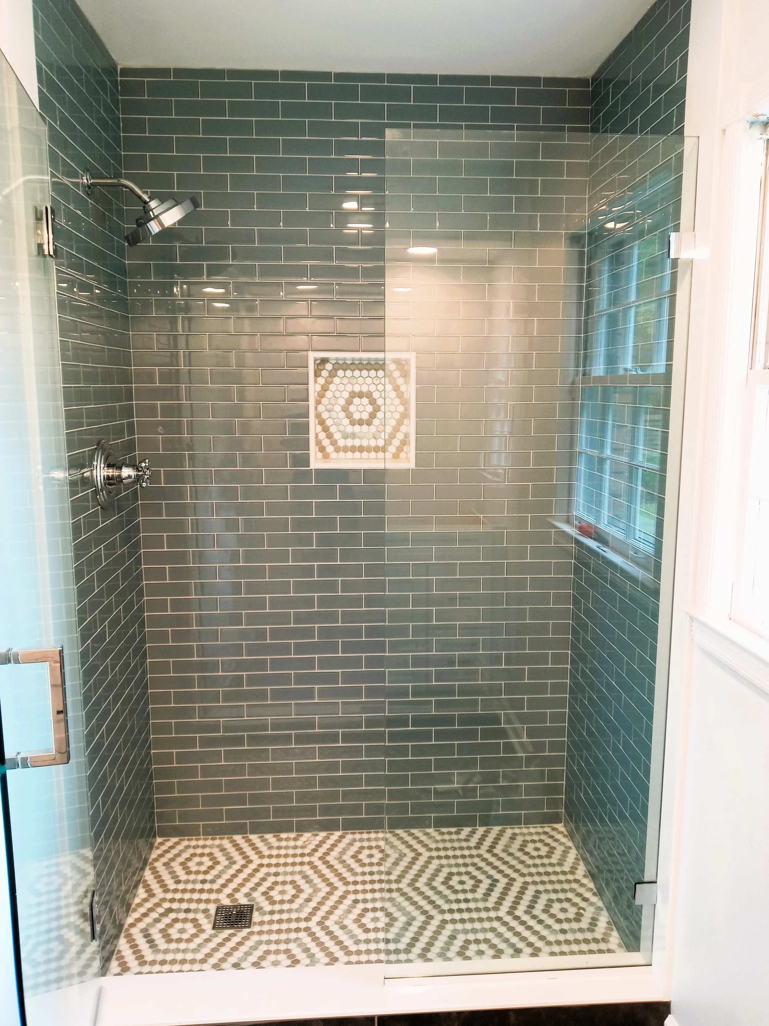 Modern glass-enclosed shower with green subway tiles on the walls and geometric patterned tiles on the shower floor, with a square niche for toiletries and a window on the right.