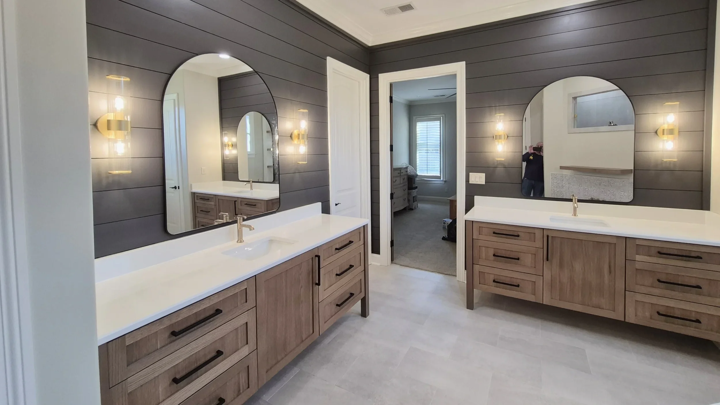 Modern bathroom with double vanity, large mirrors, wall-mounted lights, and a doorway leading to a bedroom with a window