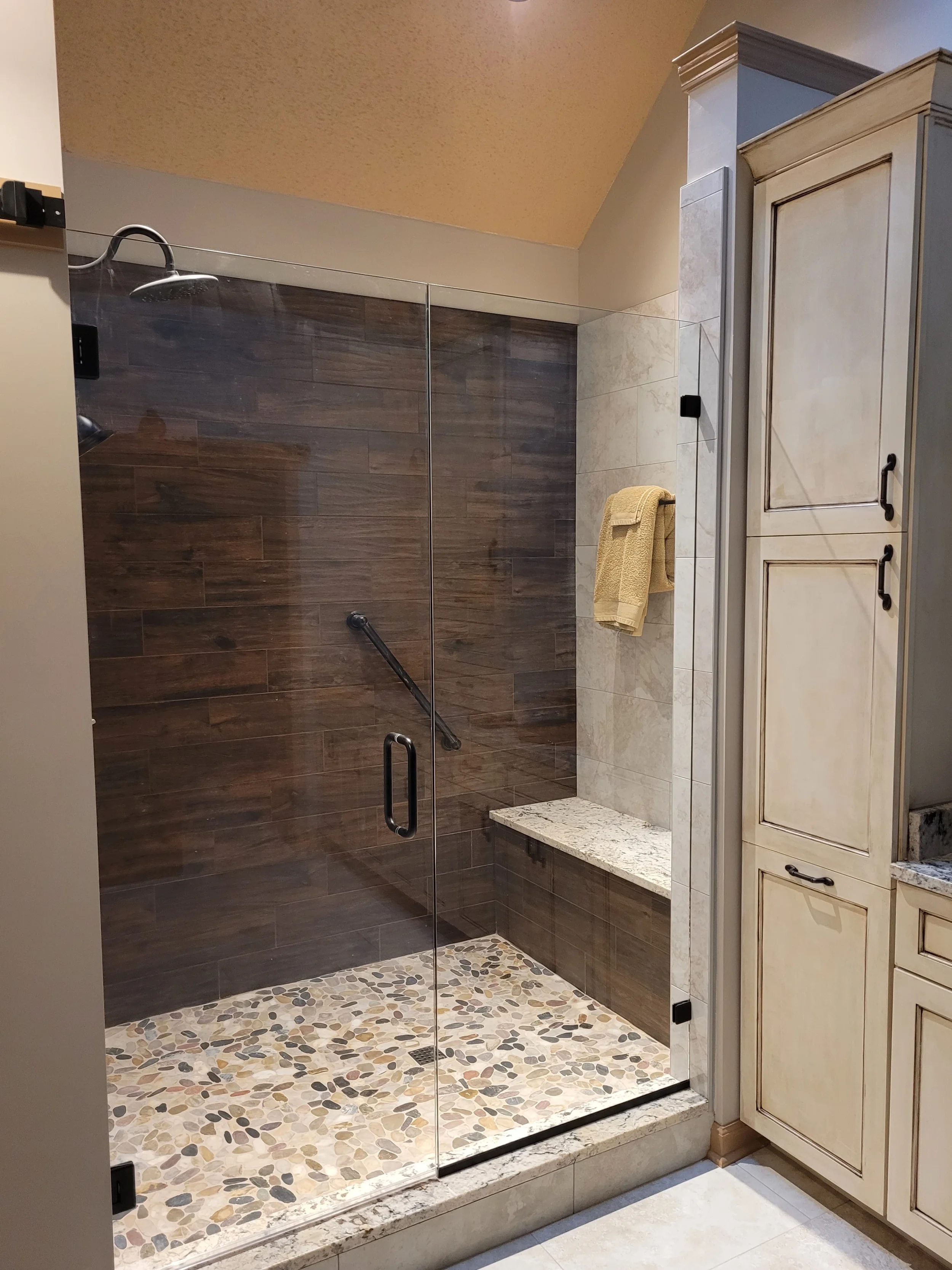 A modern walk-in shower with a glass door, pebble tiled floor, dark wood tile walls, a built-in bench, and a beige towel on a wall-mounted towel rack.