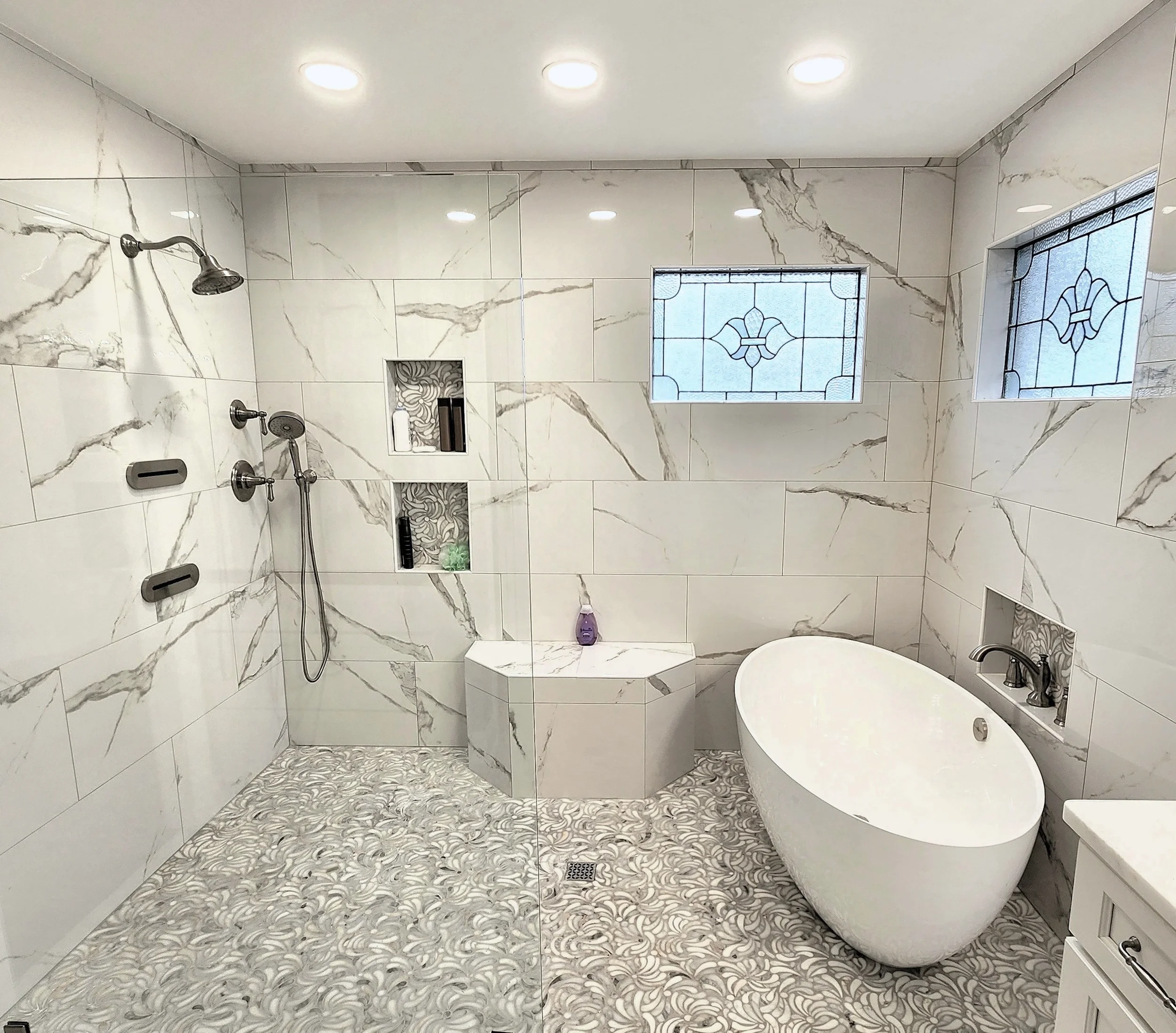 A clean glass shower stall with a chrome handle, tiled walls, and a built-in tiled seat with toiletries. It has a small niche with hexagonal tiles and a drain on the floor.