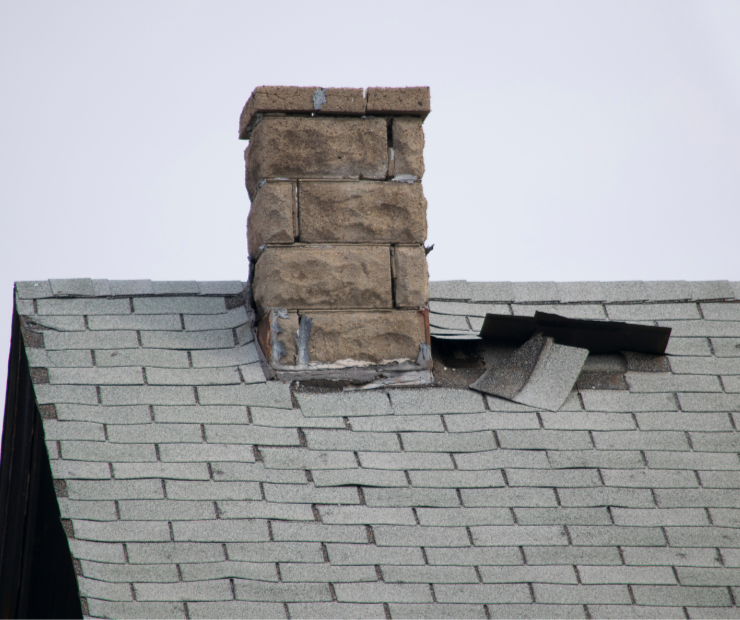 A damaged chimney on a house roof with missing and loose shingles nearby.