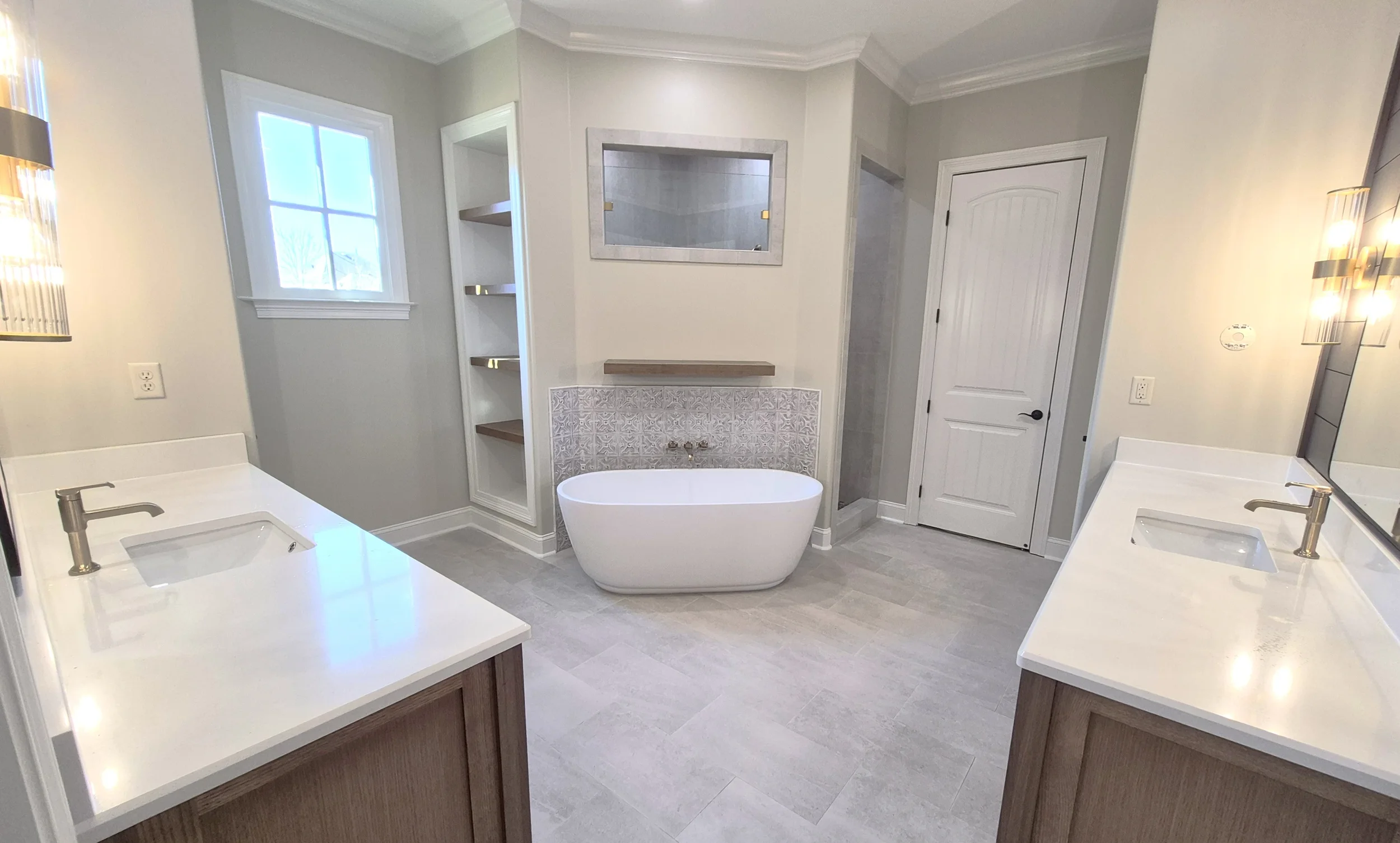 A modern bathroom with two white sinks on opposite sides, a freestanding bathtub in the center, a window, a decorative mirror, and built-in shelves.