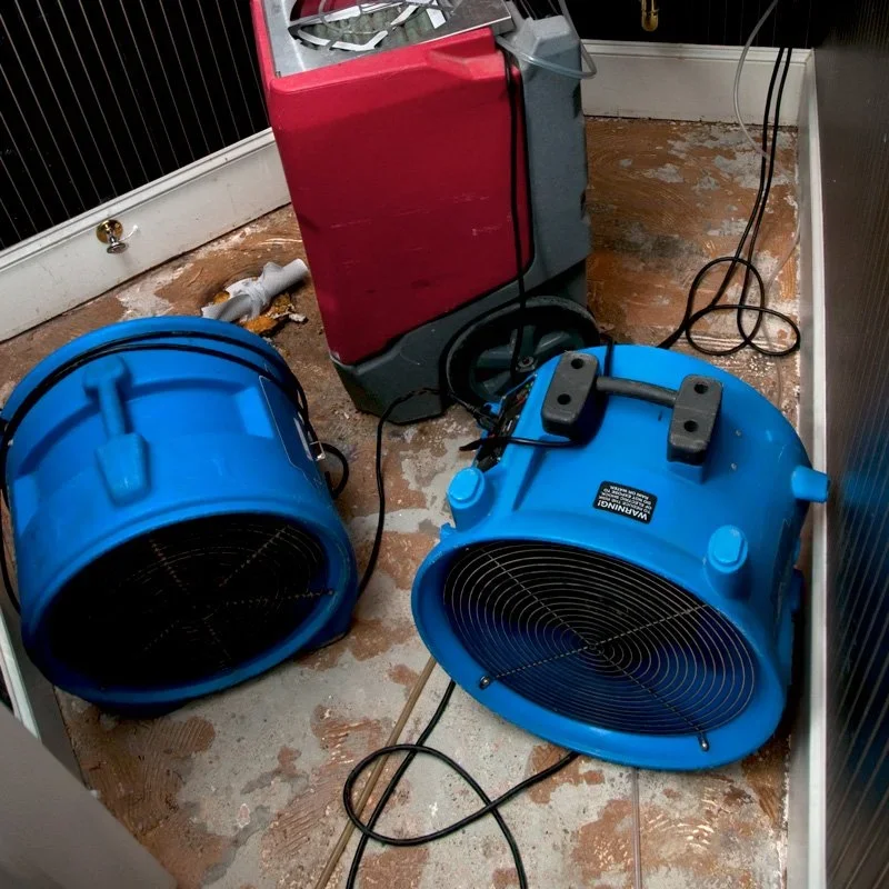 Two large blue industrial fans and a red humidifier in a small room with wooden flooring and black wall paneling drying water damage.