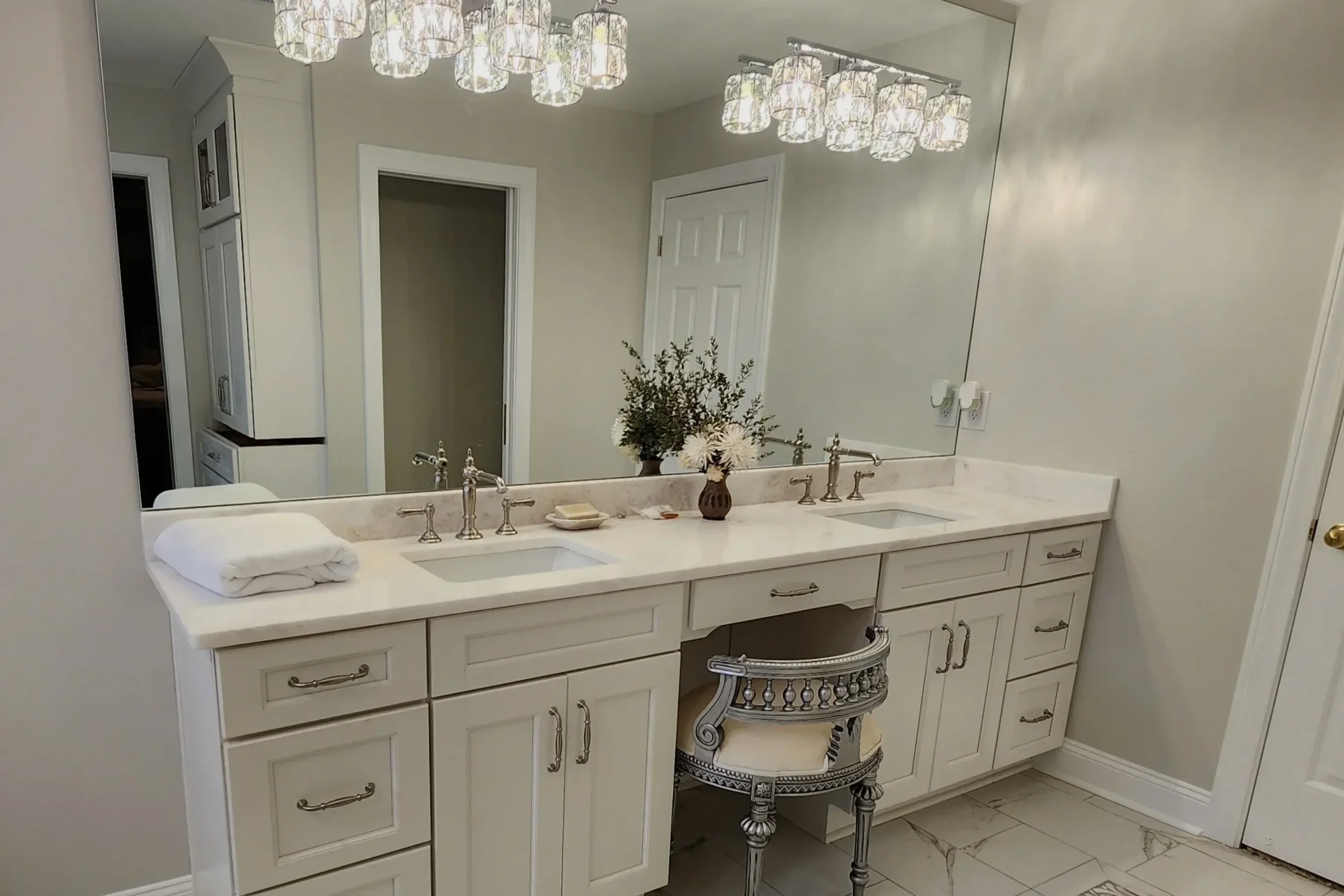 Bathroom vanity with a large mirror, white cabinetry with gold hardware, a marble countertop, two sinks, and a decorative silver stool underneath. There are two sets of ceiling lights above and a flower arrangement on the counter.