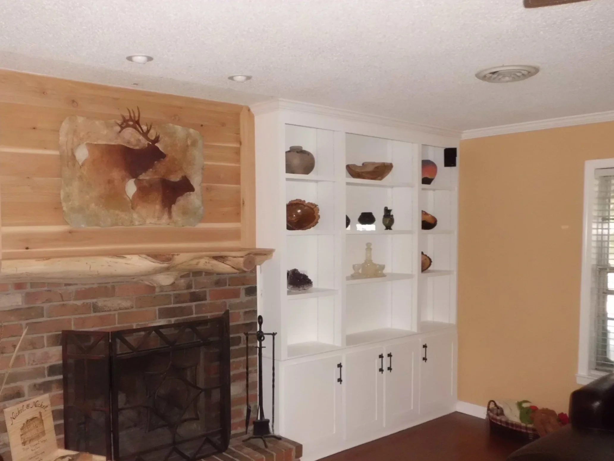 Living room with brick fireplace, wooden mantel, mounted deer artwork, white built-in shelves with decorative objects, and a leather sofa.