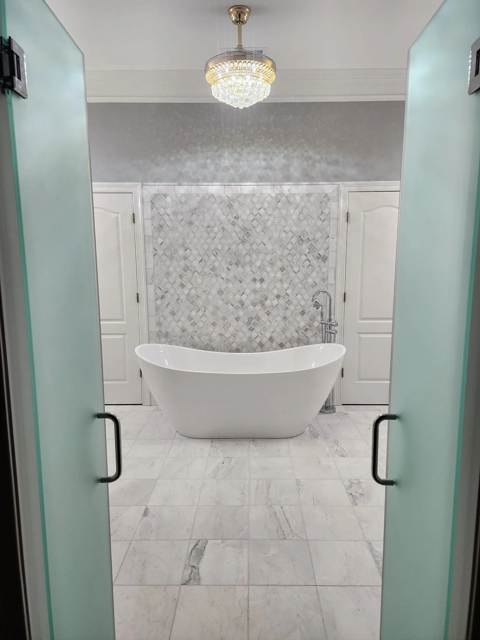 Modern bathroom with a standalone white bathtub, a textured gray tile wall, marble floors, white doors, a chandelier, and frosted glass double doors.