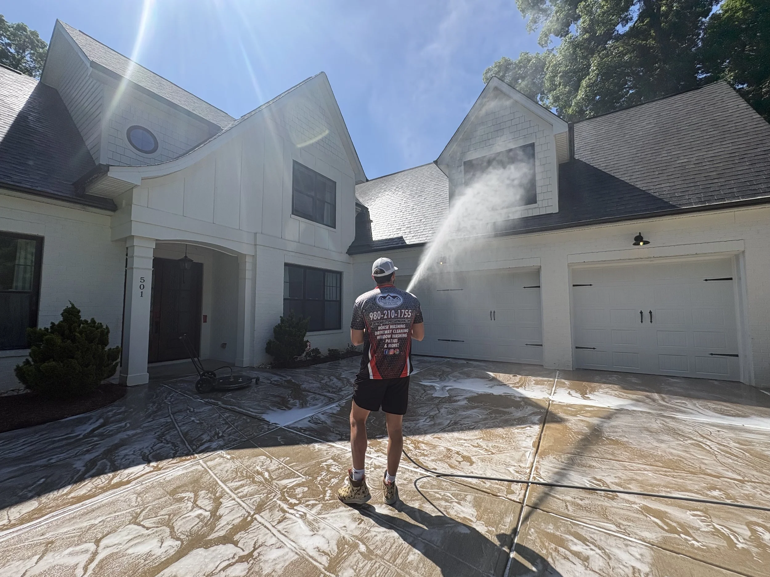 Person using pressure washer to clean the driveway in front of a white house on a sunny day.