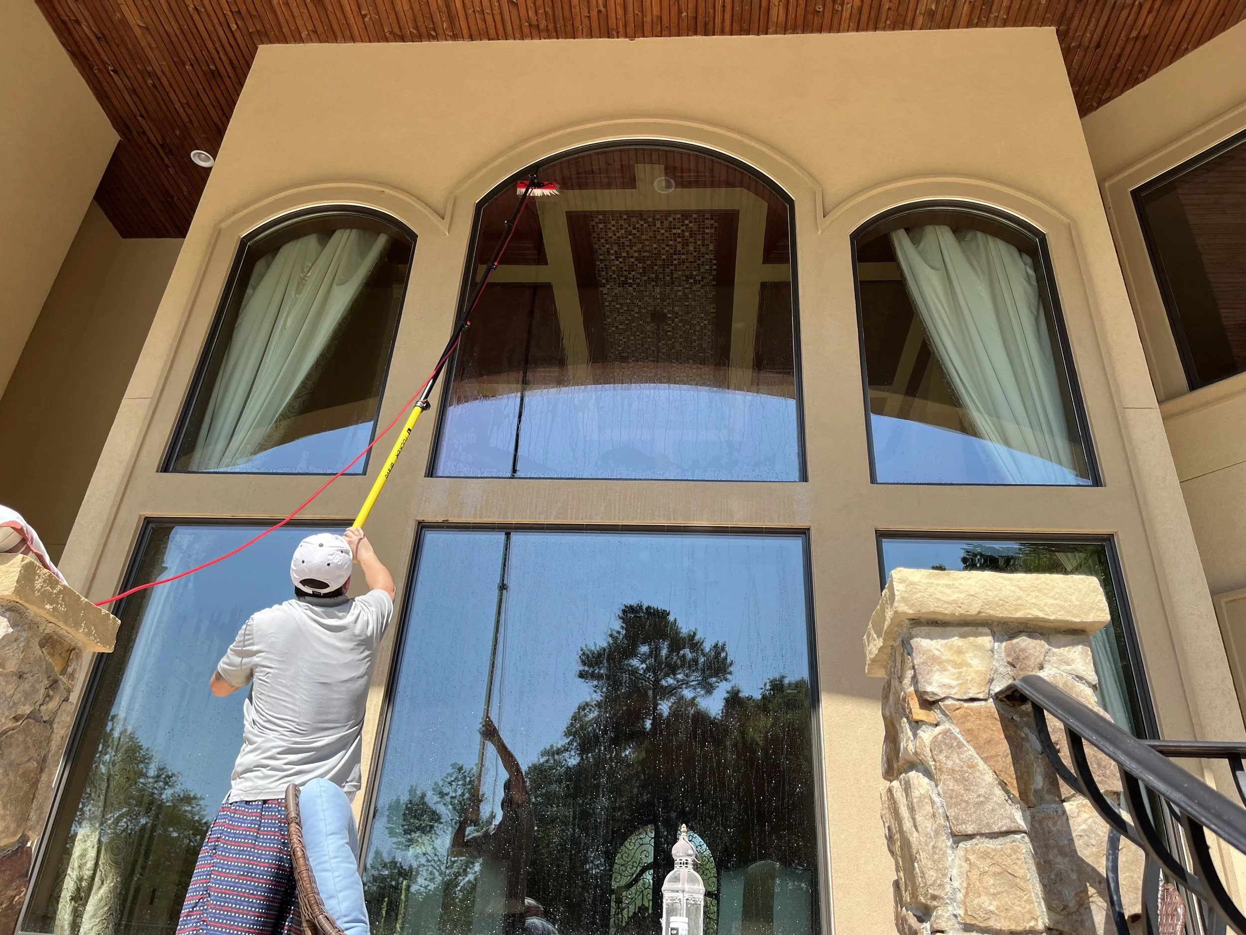 A person in a gray shirt and cap uses a pole to clean a large arched glass window of a house with stone and stucco exterior walls.