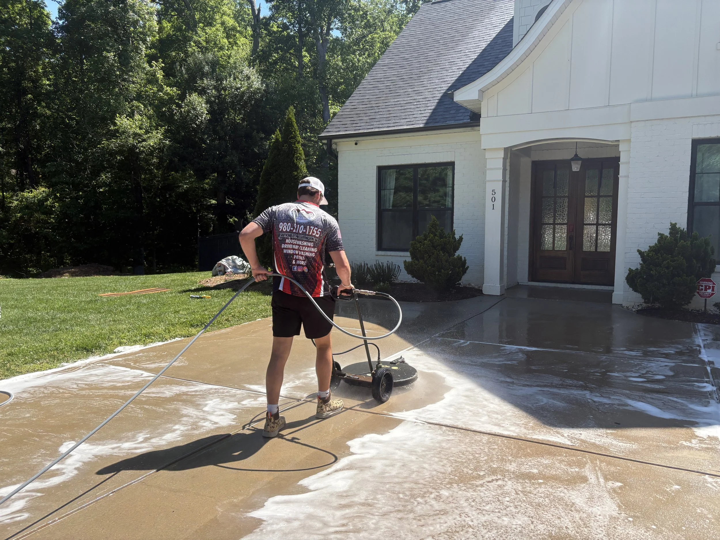 A man watering a backyard lawn with a hose during daytime, in front of a white house with a dark front door and large windows, trees in the background, and a warning sign on the lawn.