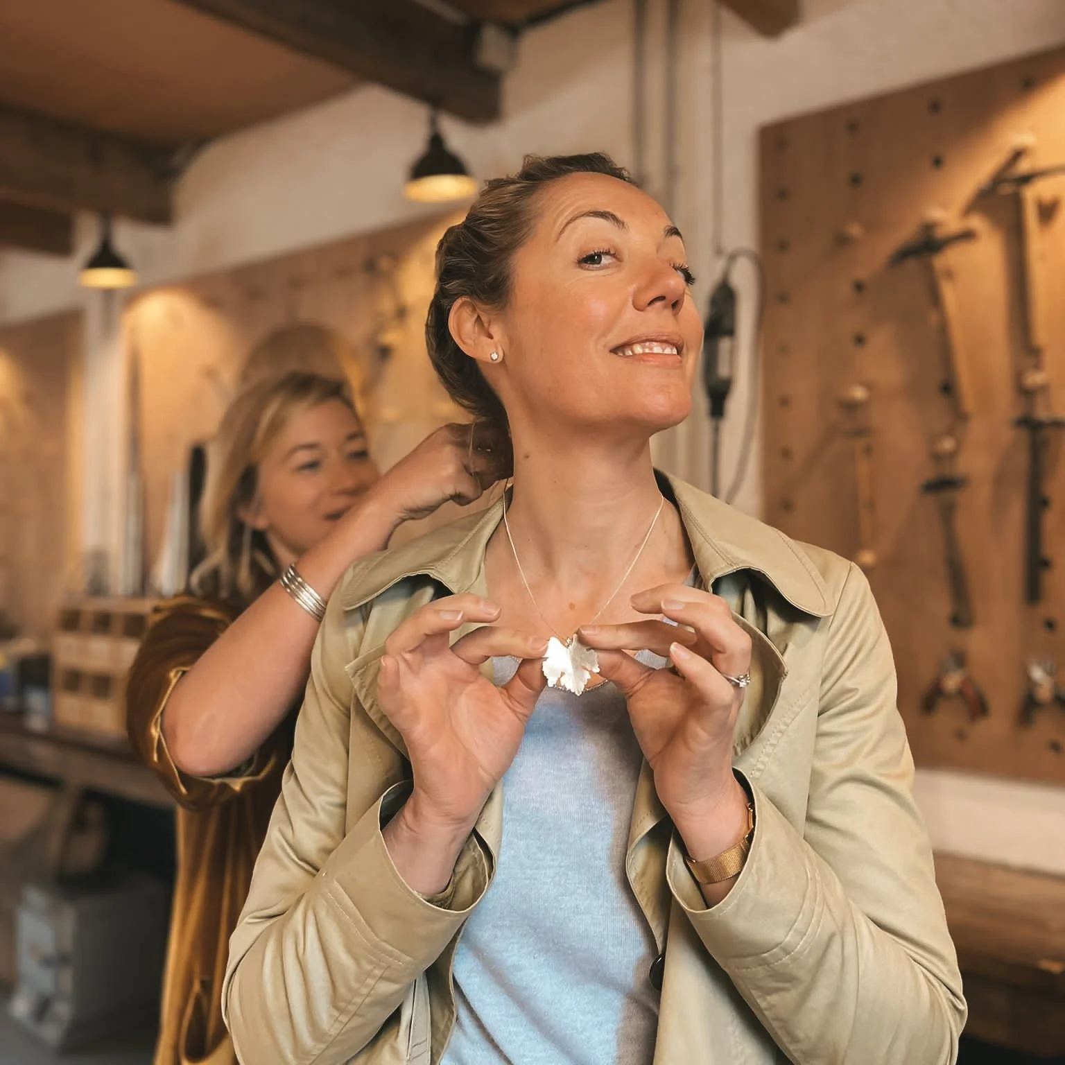 Two women in a workshop, one helping the other put on a necklace, with tools hung on the wall in the background.