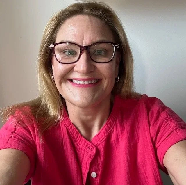 A woman with light hair, glasses, and earrings smiling while taking a selfie. She is wearing a pink top, and the background is plain white.