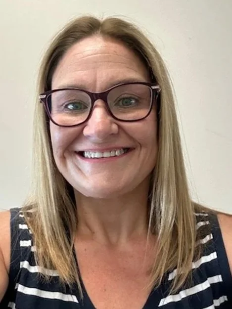 A woman with shoulder-length blonde hair, wearing glasses and a black and white striped sleeveless top, smiling at the camera against a plain background.