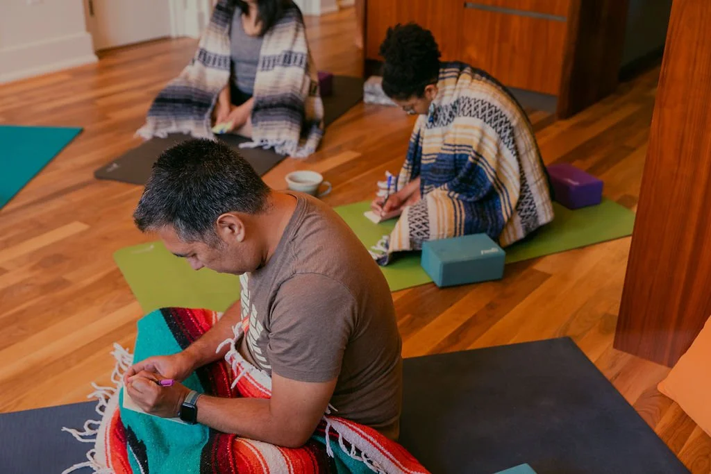 Three people sitting on yoga mats in a room, engaged in a mindful activity or meditation session. The room has wooden floors and a cozy atmosphere.