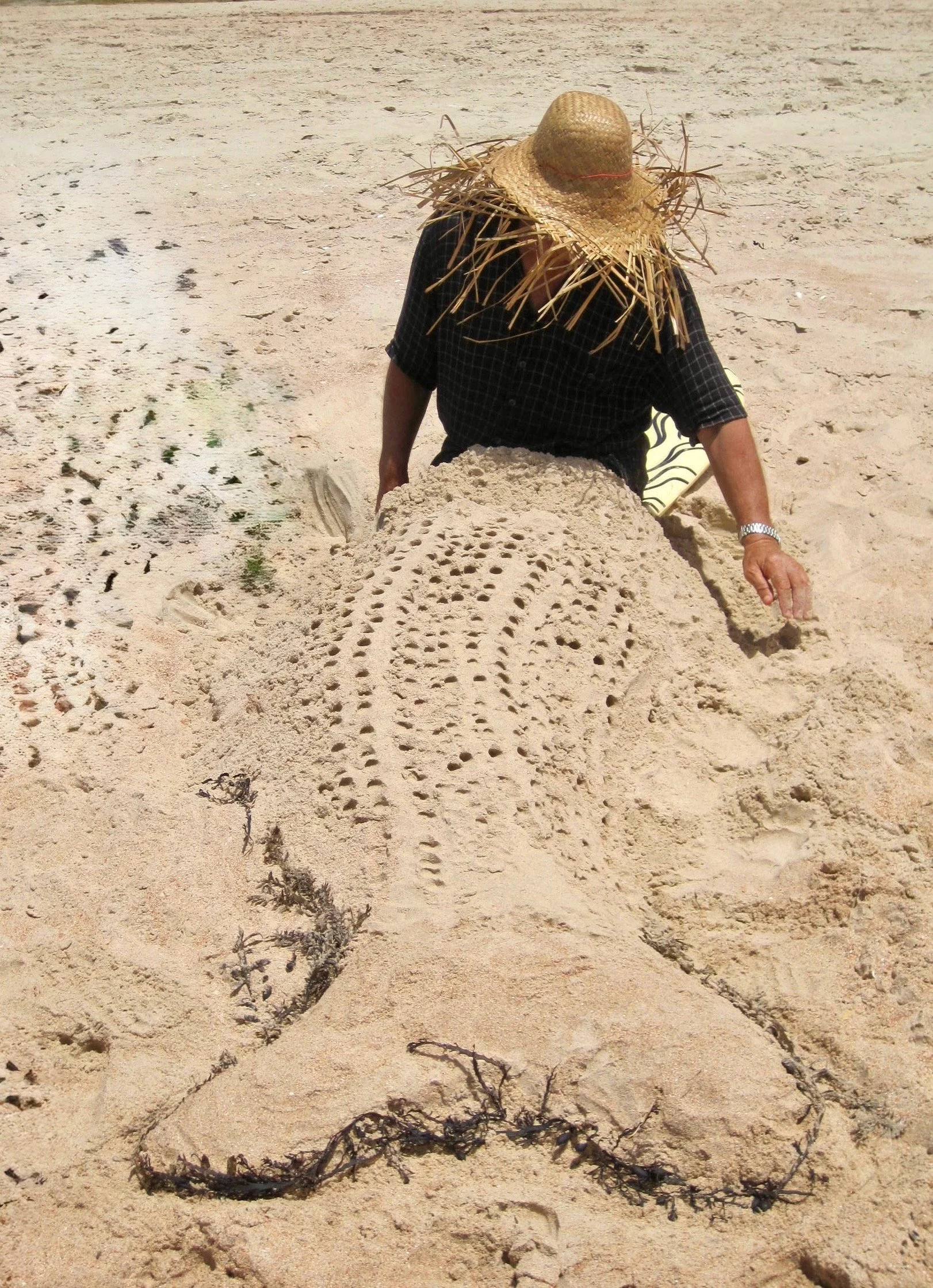 Person in a sun hat sitting alone on the sand at the beach, seen from behind