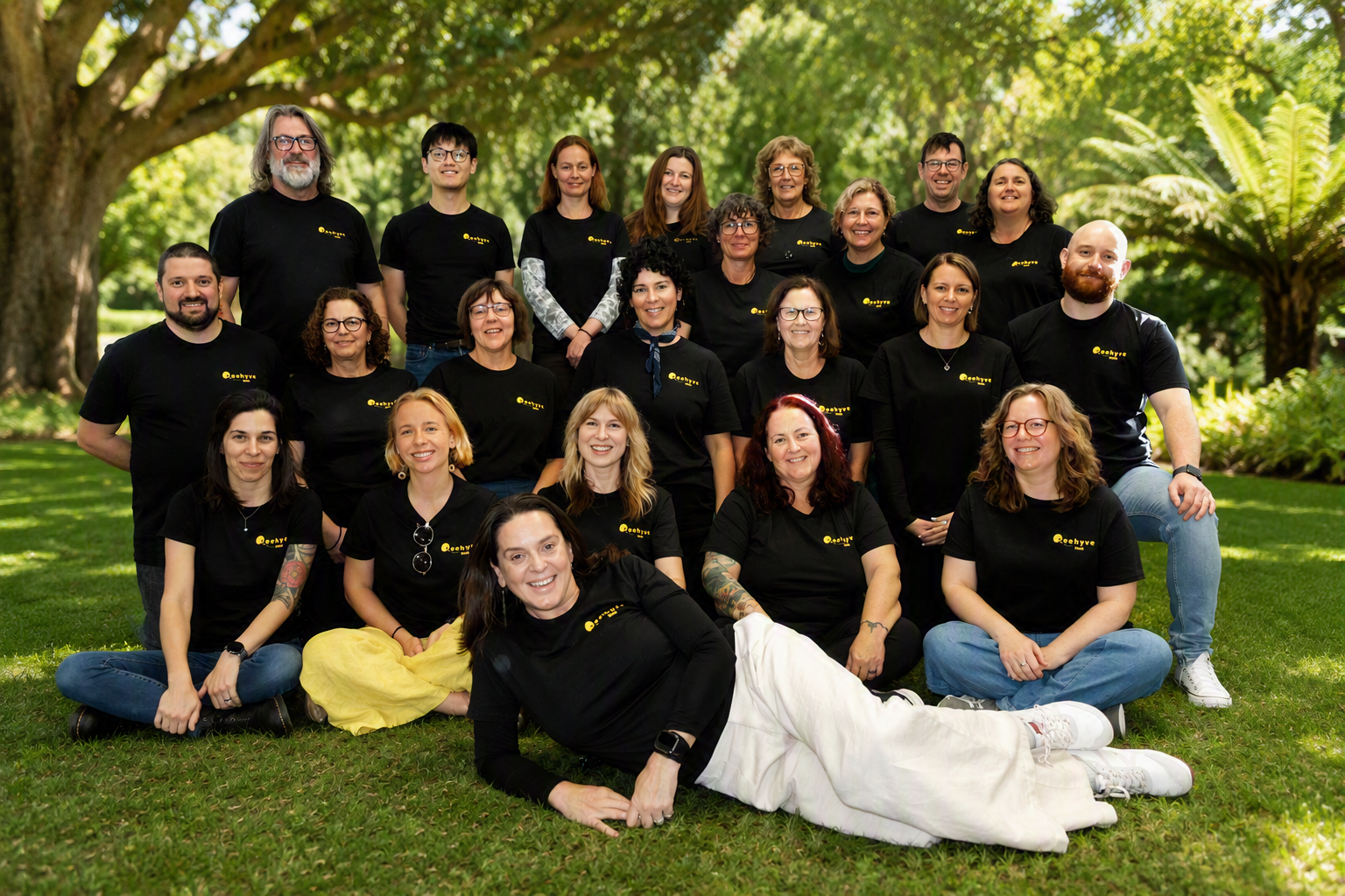 Group of diverse people wearing black t-shirts with yellow logo, posing outdoors on green grass with large trees in the background, some sitting, some standing, and one lying in front.
