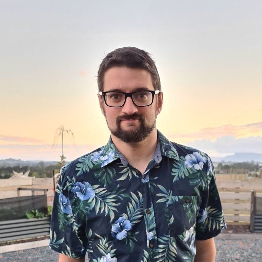 A man with glasses and a beard, wearing a Hawaiian shirt with blue flowers and green leaves, stands outdoors during sunset with a calm landscape in the background.