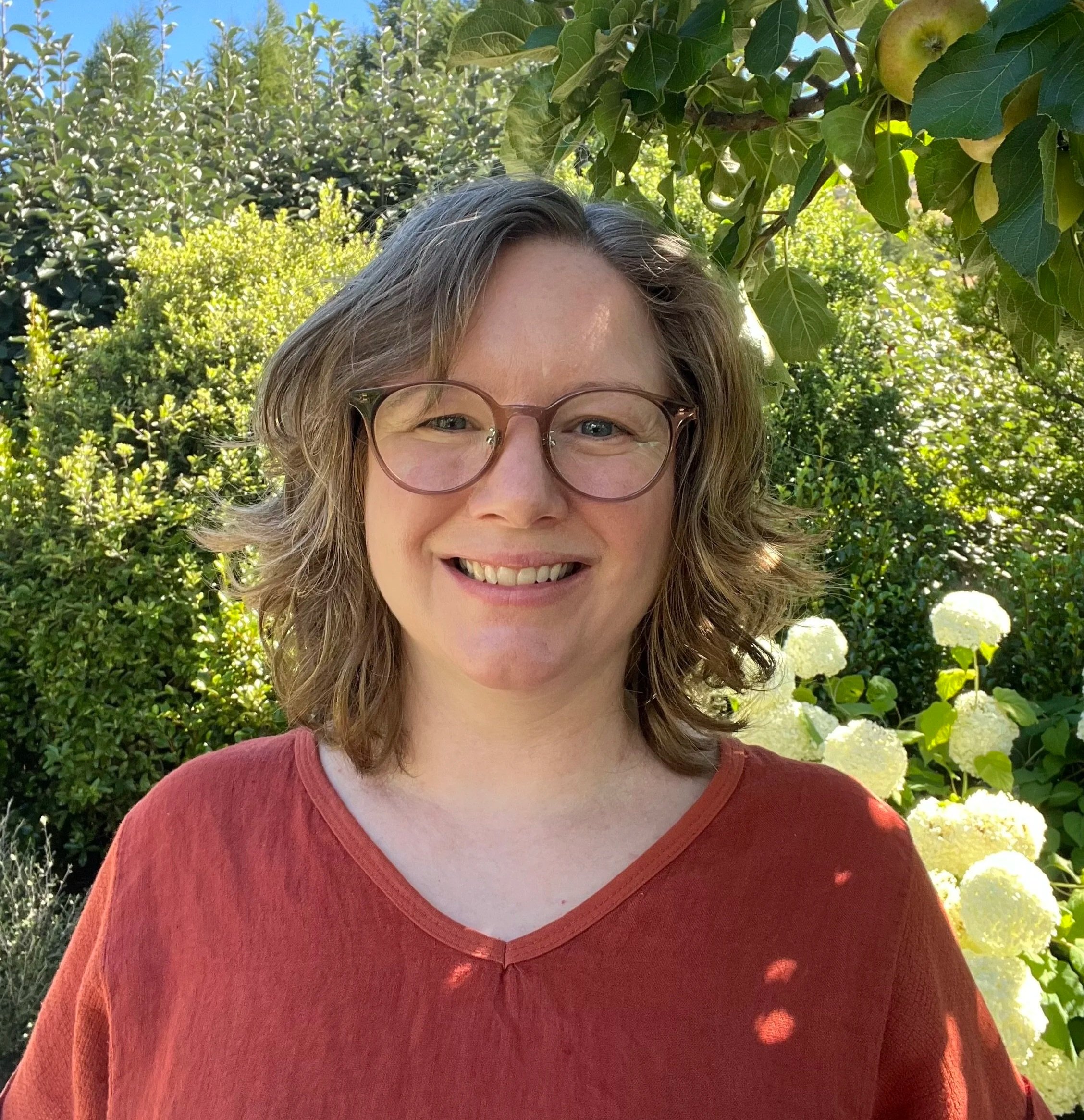 Close-up of a woman with glasses smiling outdoors with green foliage and white flowers in the background.