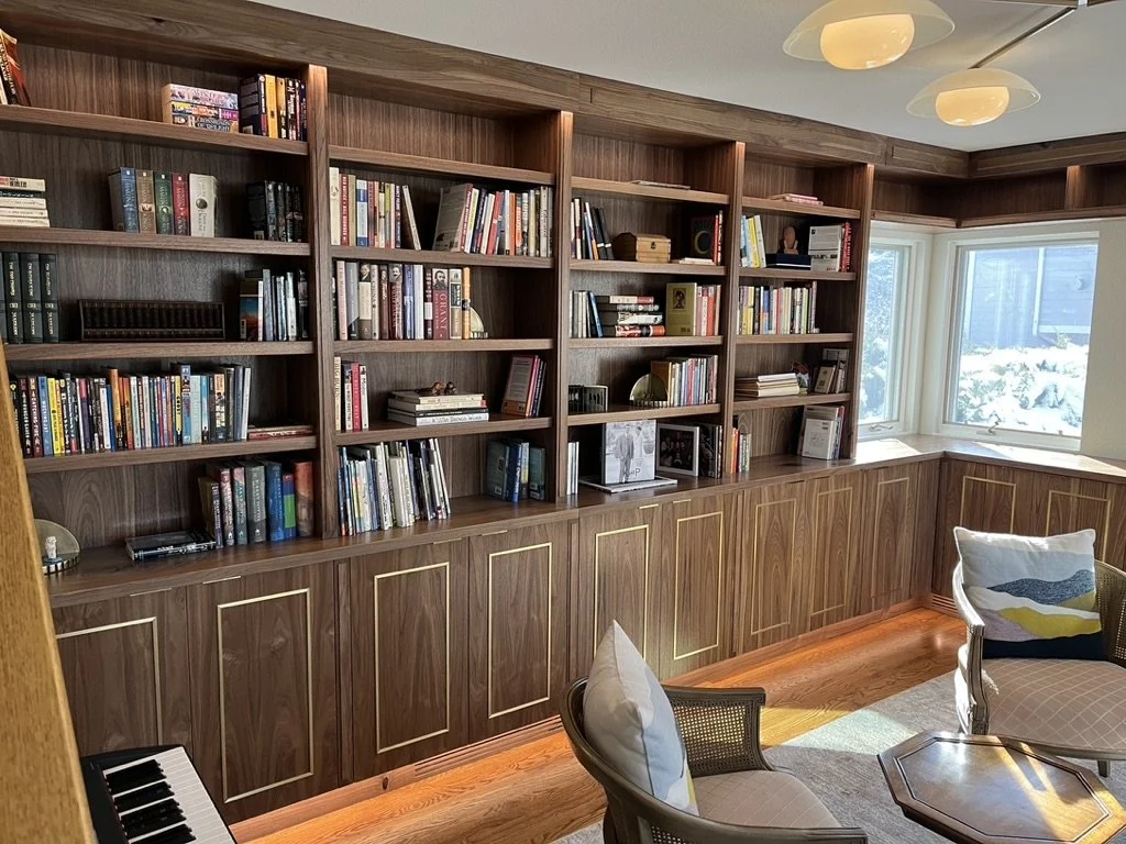 A wooden bookshelf covering the wall with books and decorative items, beside a window with snow outside. There are two chairs and a piano in the room.