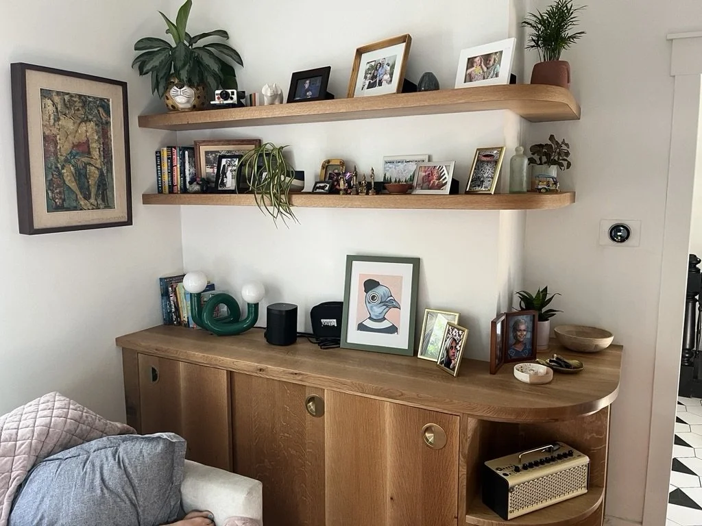 Wooden sideboard with framed photographs, decorative objects, plants, and a speaker, with two wooden wall shelves holding framed photos, books, and decorative items, over a white wall.
