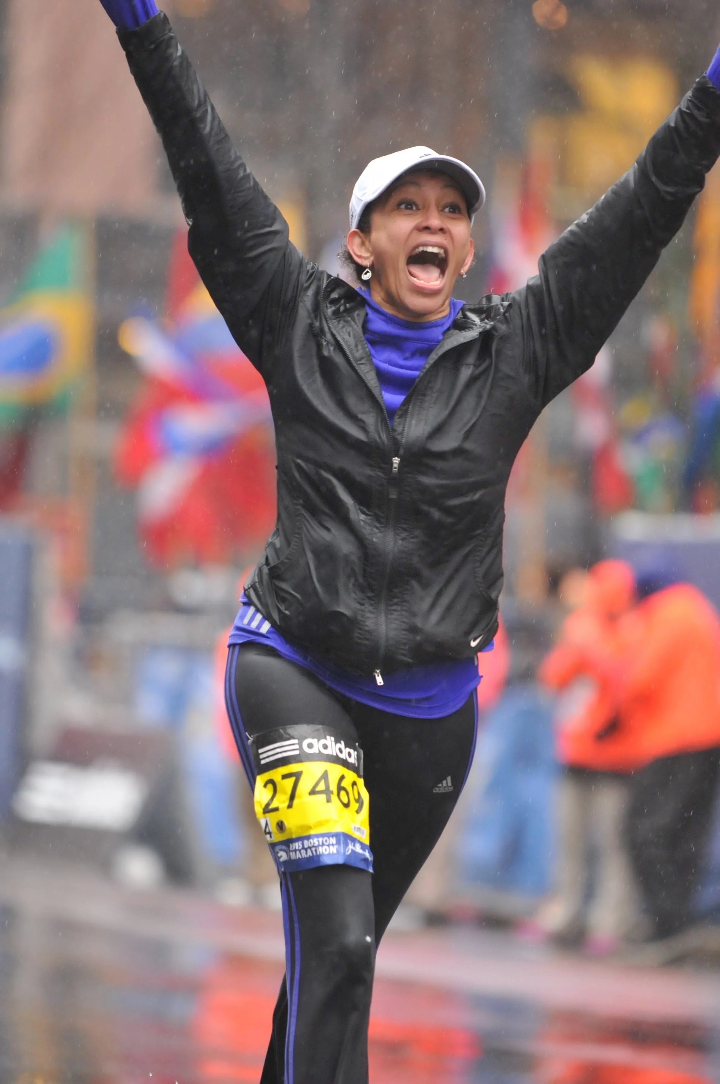 Jen Kanygui in running rain gear at the Boston Marathon. The rain is coming down and she has her arms lifted, shouting for joy.