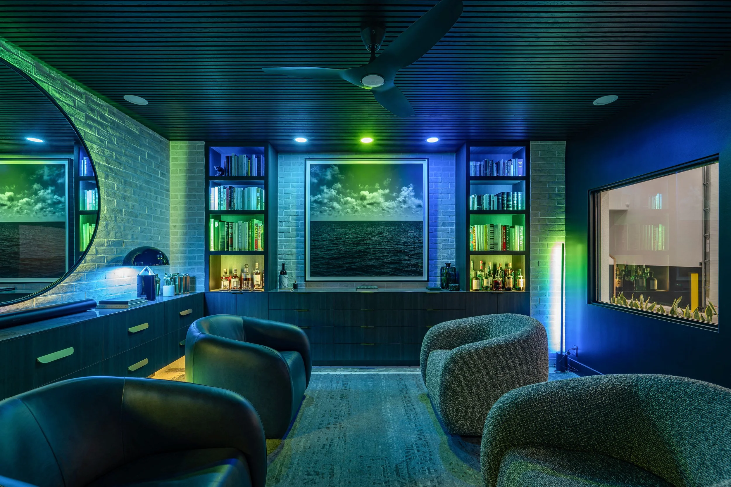 Modern lounge room with ambient lighting, featuring a ceiling fan, large circular mirror, built-in shelves with books and bottles, a central ocean-themed artwork, and contemporary chairs.