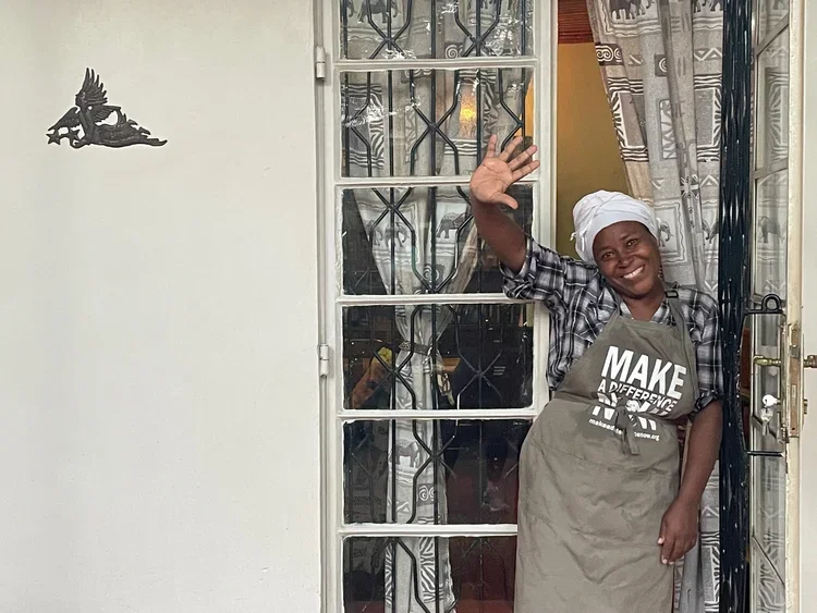 A smiling woman in a headscarf and apron waves through a glass door, standing in a doorway with patterned curtains and decorative ironwork.