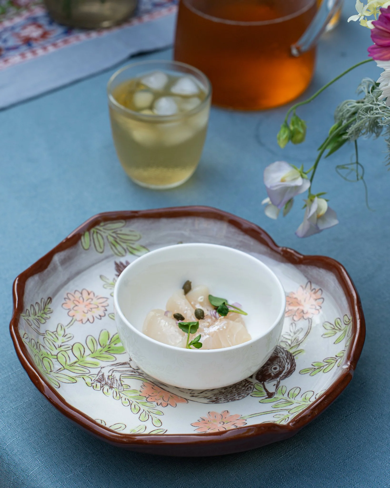 A bowl of colorful sashimi topped with microgreens, crispy onions, and salmon roe is placed on a marble countertop. Behind the bowl, there is a white rectangular dish holding wooden chopsticks. Part of a printed menu is visible in the lower right corner of the image.