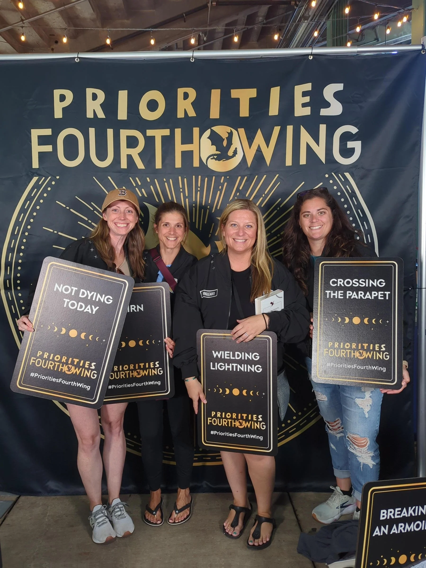Four women posing together at an event in front of a backdrop that reads 'Priorities Fourth Wing.' They are holding signs with phrases like 'Not Dying Today,' 'Wielding Lightning,' and 'Crossing the Parapet,' related to the book 'Priorities Fourth Wing.'