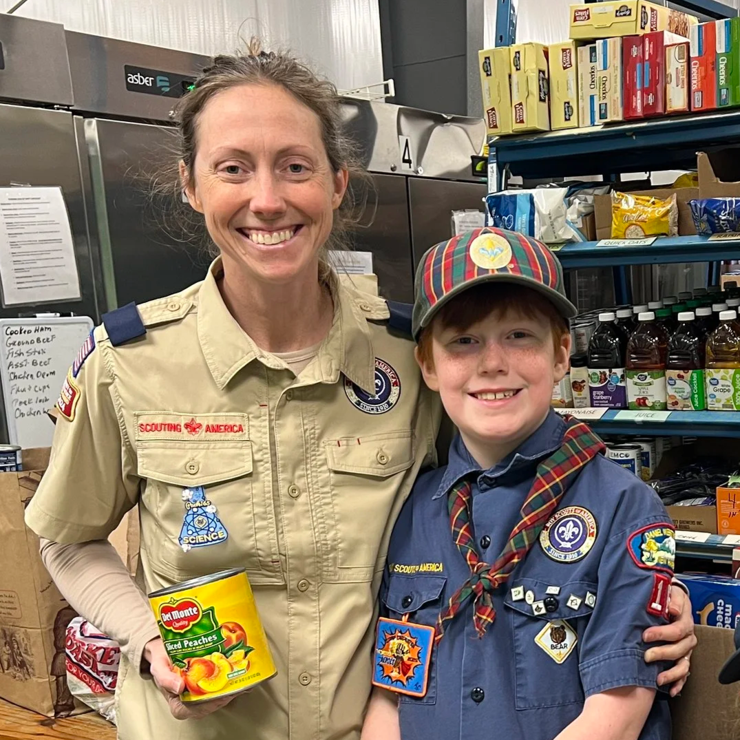 A woman and a boy dressed in Boy Scout uniforms, smiling inside a store with shelves of snacks and bottled drinks in the background. The woman is holding a can of Del Monte peach slices.