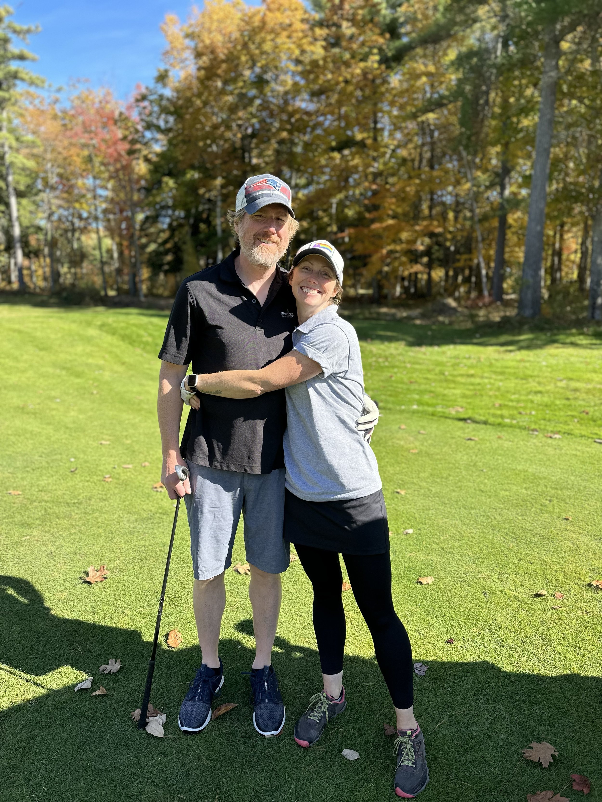 A man and woman hugging on a golf course during autumn, with trees having colorful fall foliage in the background.