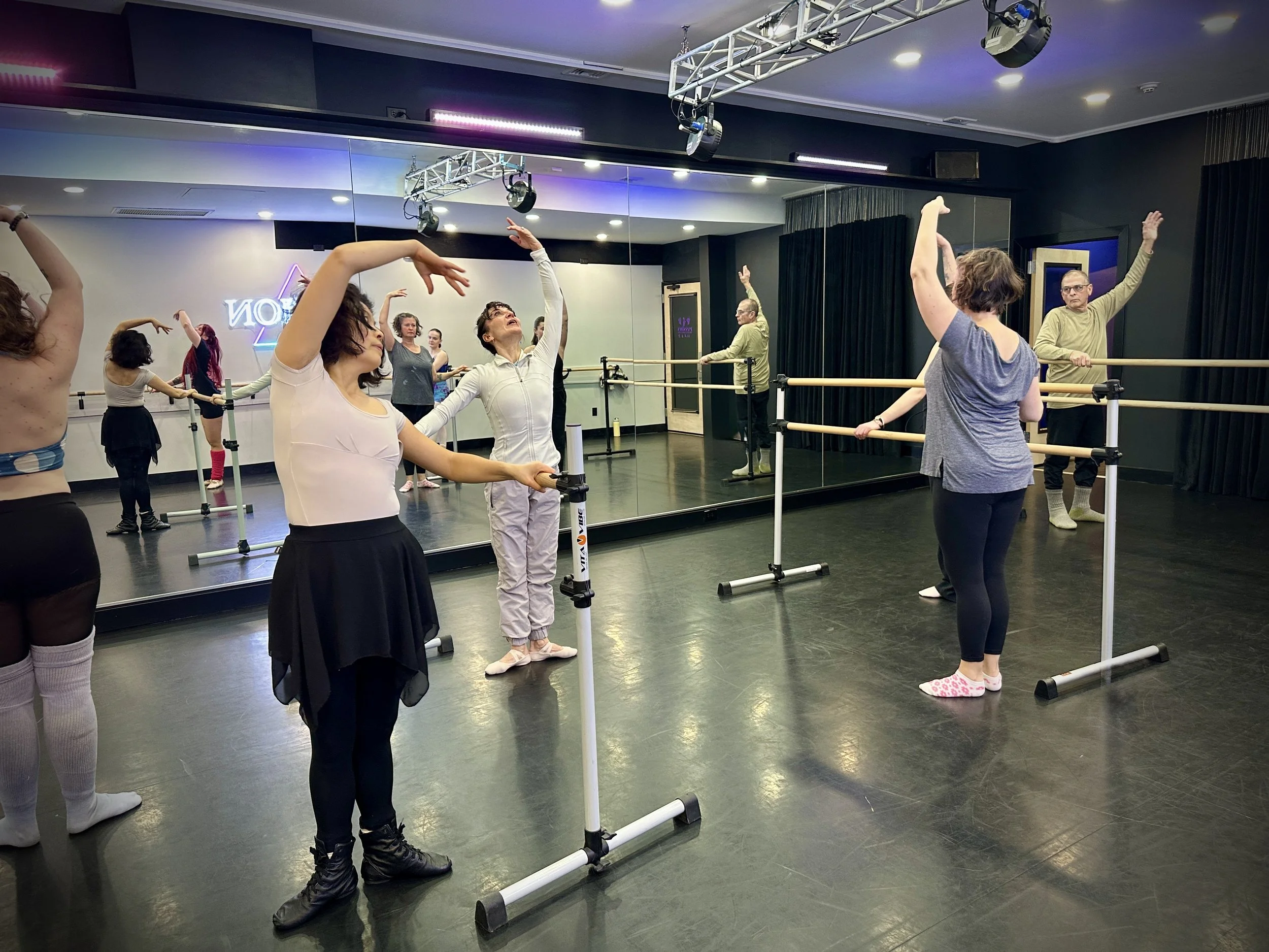 A group of people in a dance class performing ballet exercises at a barre in a blue room with wooden flooring.