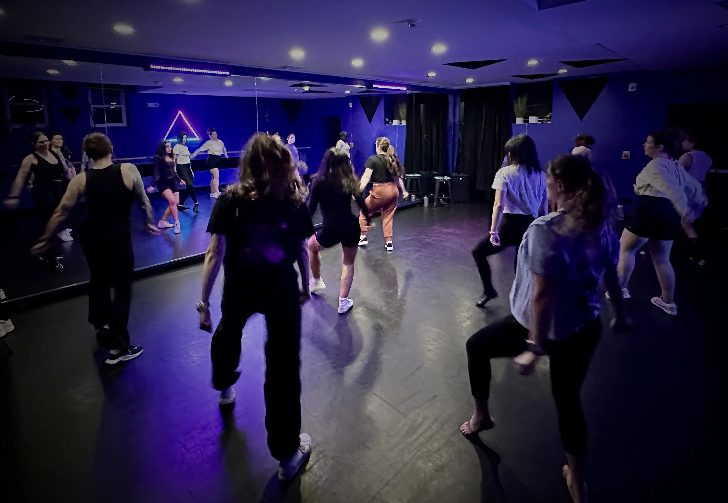 Five women in black outfits posing in front of a blue wall with a ballet barre, standing on a wooden floor.