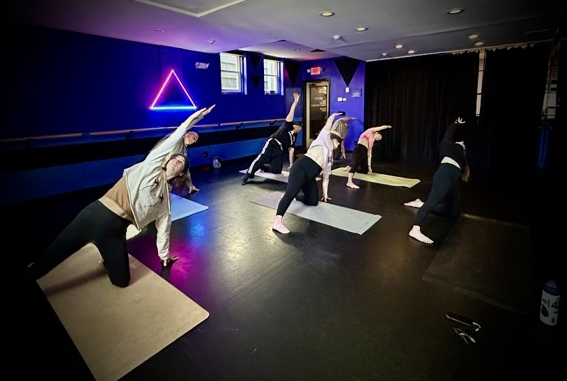 A woman is performing a yoga pose on the floor, stretching forward with her arms extended and her legs crossed behind her, looking up at the camera with a smile.