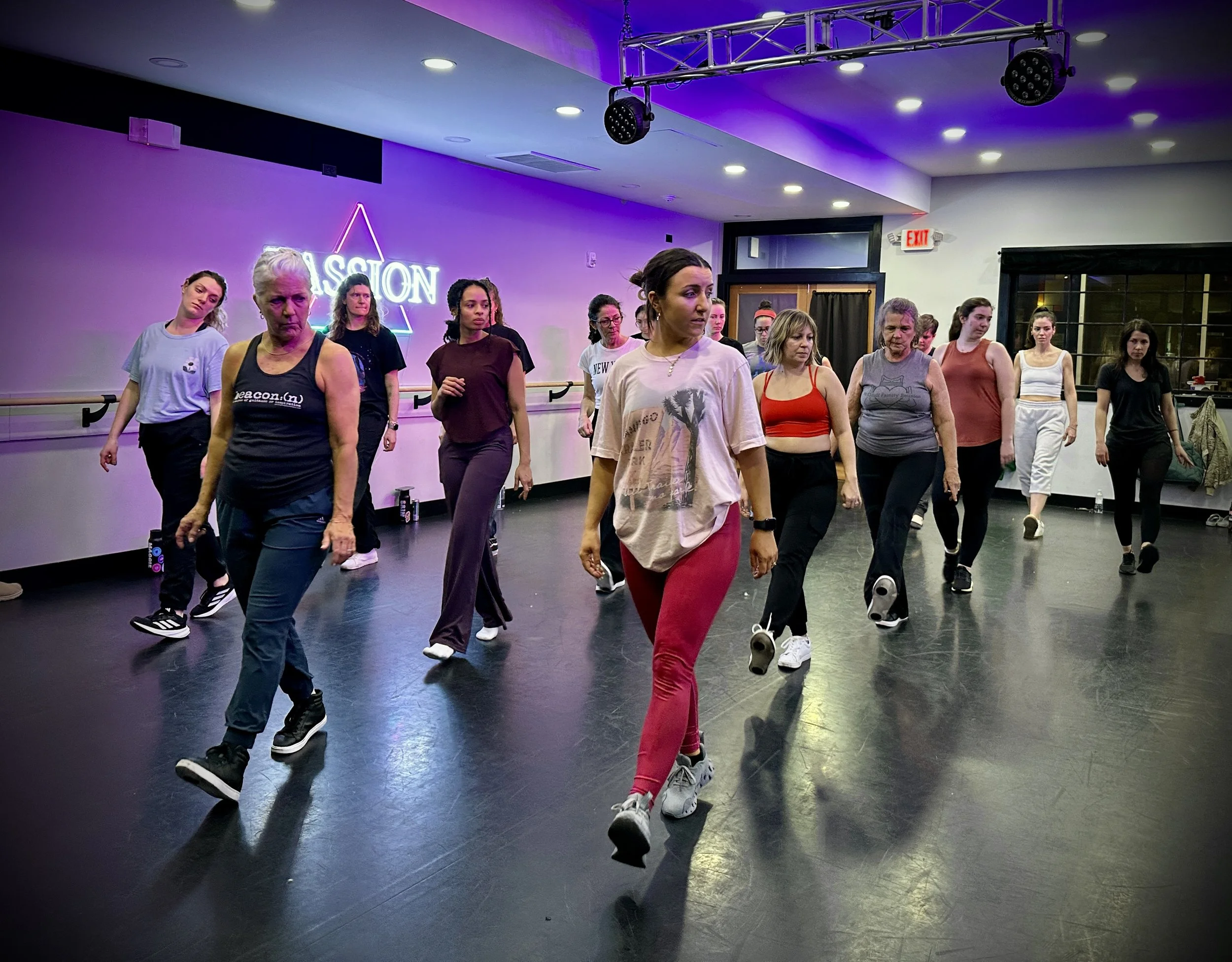 Group of people in a dance class performing a choreographed routine in a studio with purple walls and wooden floors.