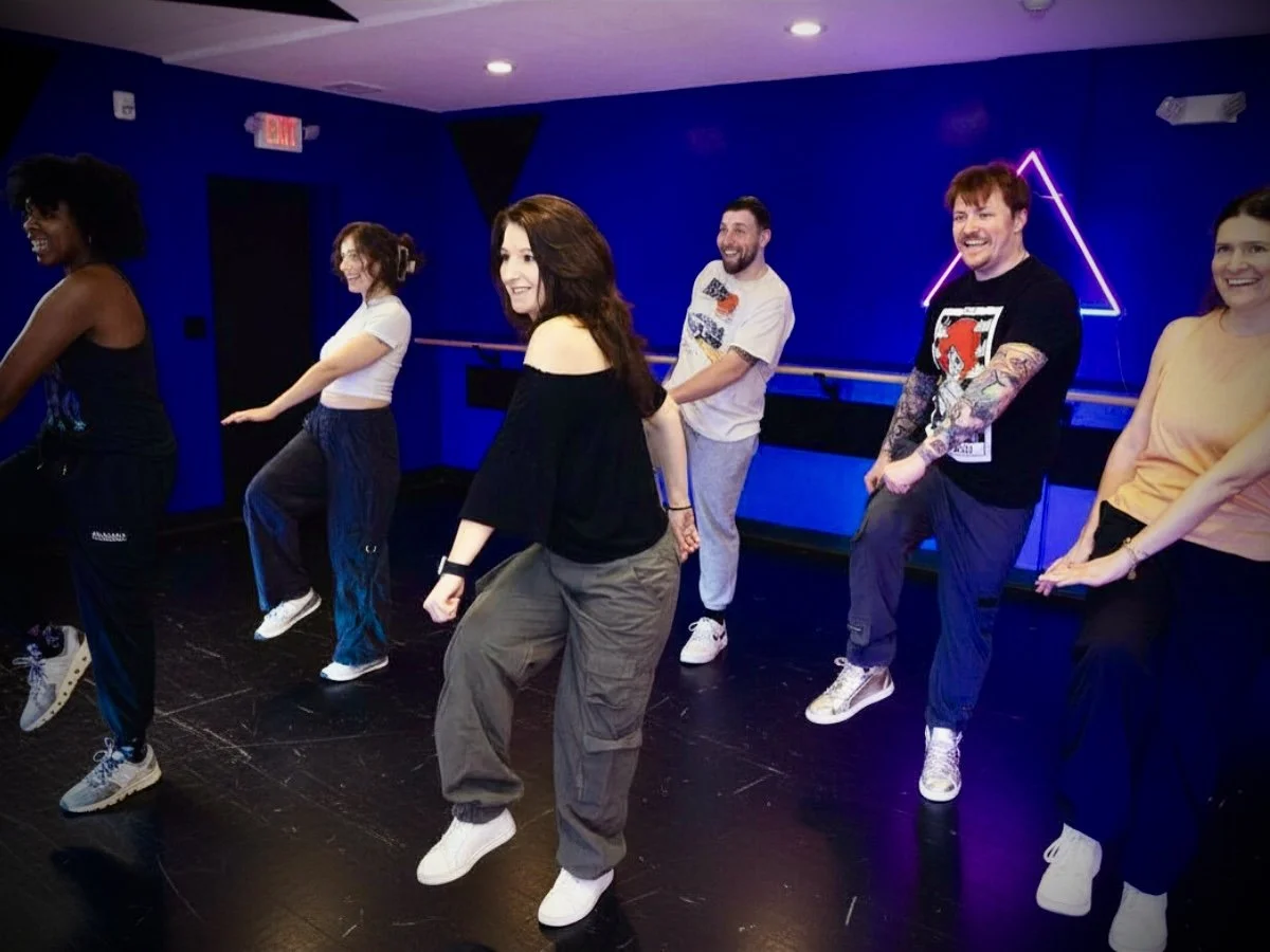 A group of six people dancing in a studio with blue walls, wooden floor, and a purple neon triangle on the wall. They are smiling and wearing casual clothing.