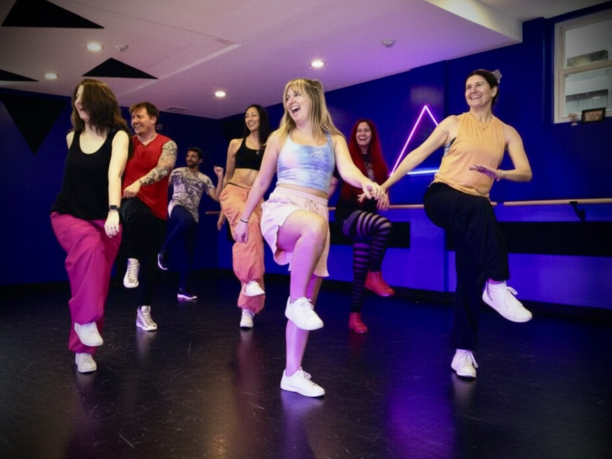 A group of men and women dancing energetically in a brightly lit studio with wooden floors and blue walls. They are wearing casual dance attire and appear to be having fun.
