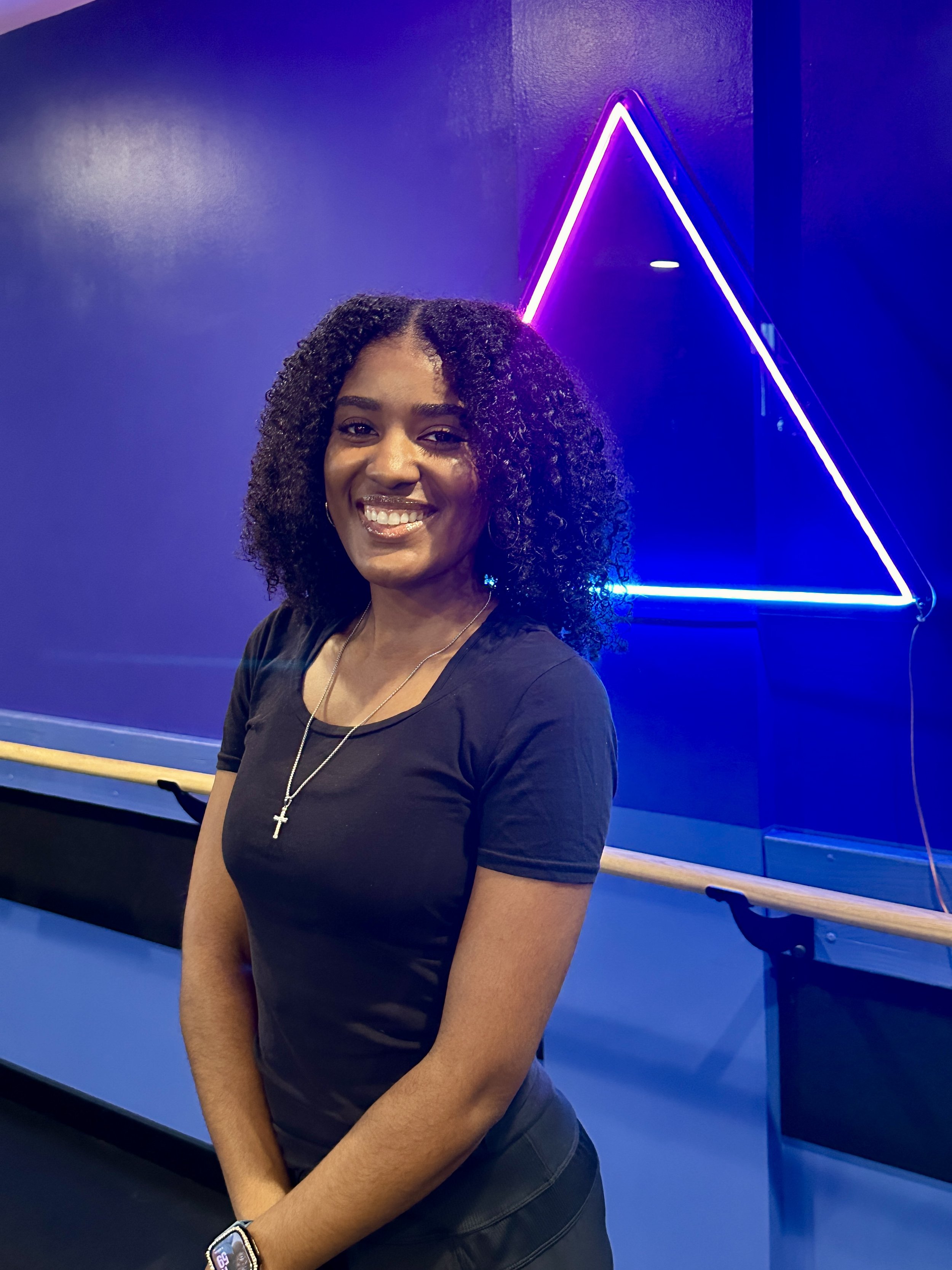 Smiling African American woman with curly hair, wearing a denim jacket and white shirt, against a pink background.