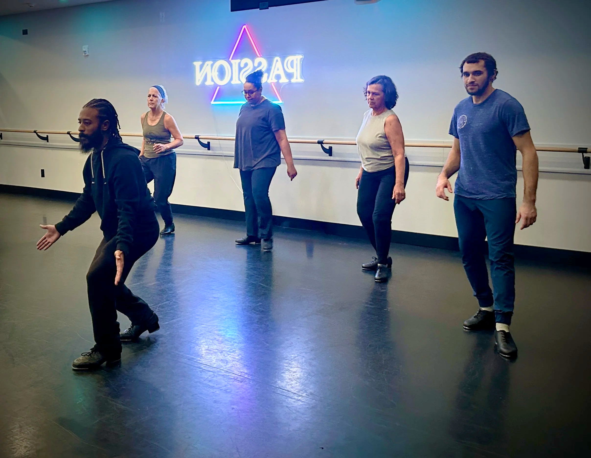 A man with a beard and braided hair smiling in front of neon triangle light on a blue wall.