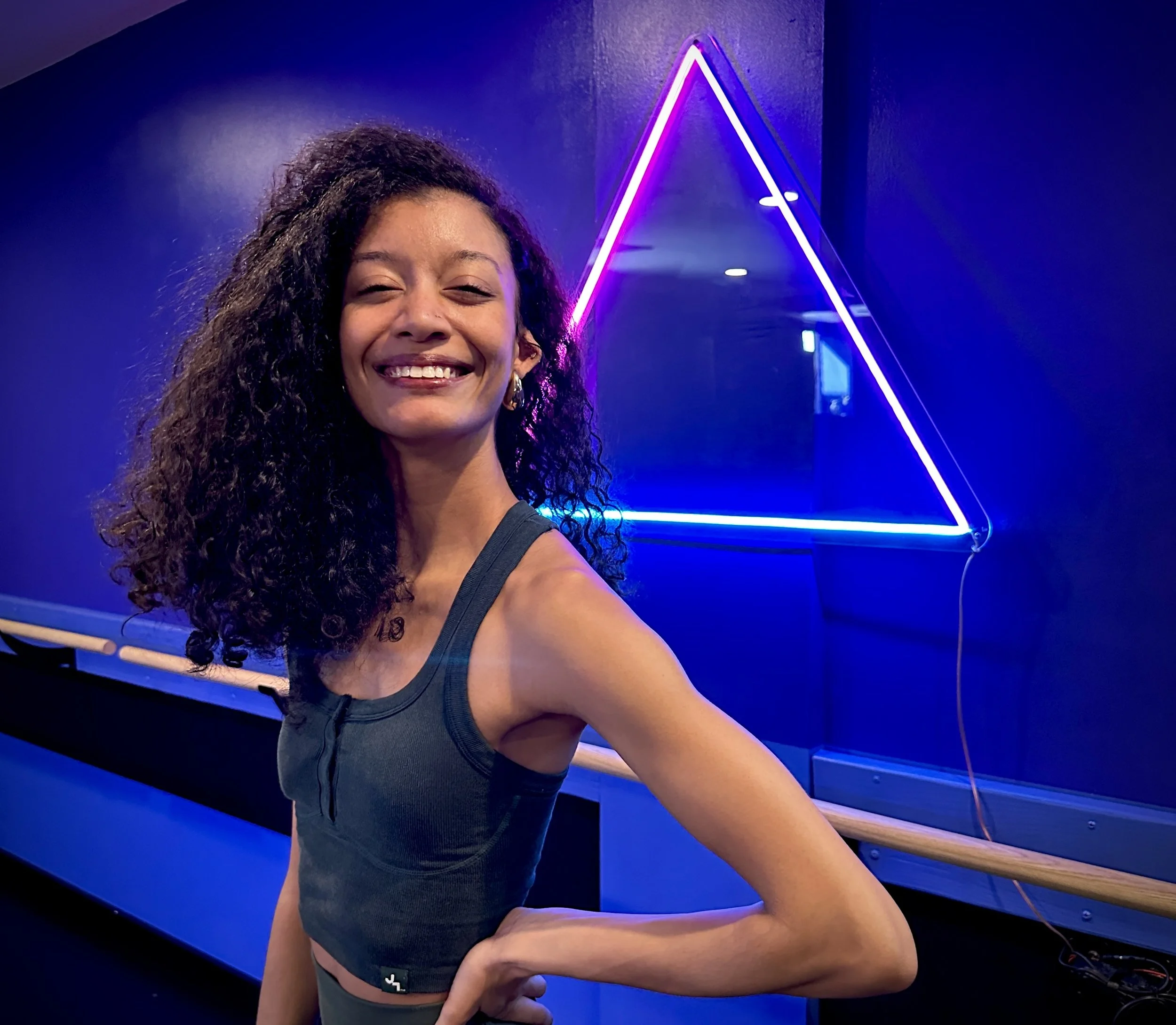 Smiling woman wearing a blue beaded dress with a keyhole cutout, black hair styled short, and a blue hair accessory, with a man in a black shirt in the background