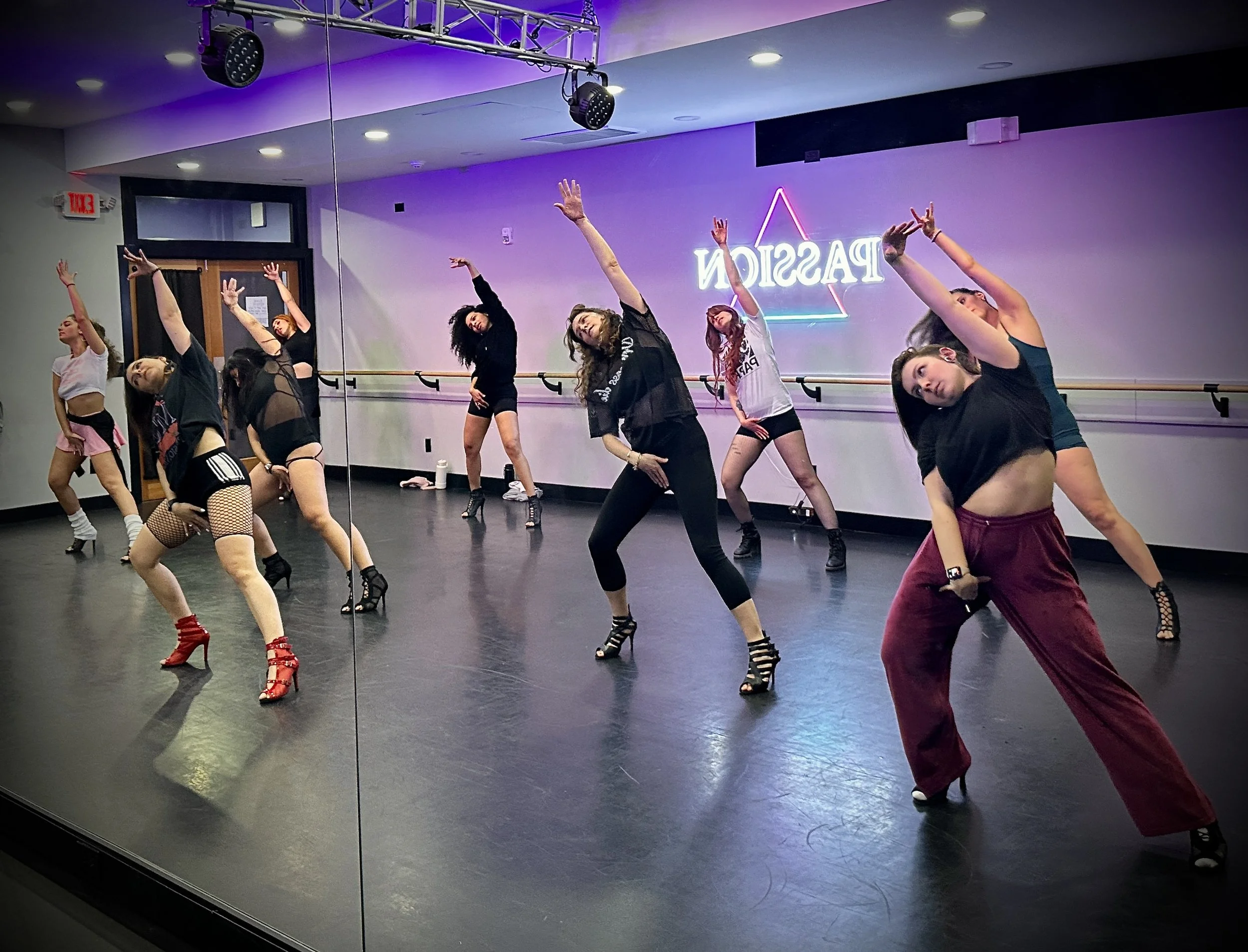 Group of women dancing in a studio with wooden floors, wearing high heels and form-fitting outfits, with blue walls and neon lights in the background.