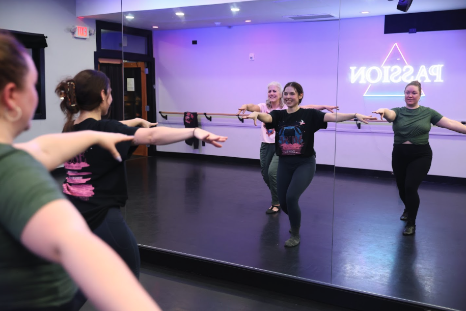 Five women in black outfits posing in front of a blue wall with a ballet barre, standing on a wooden floor.