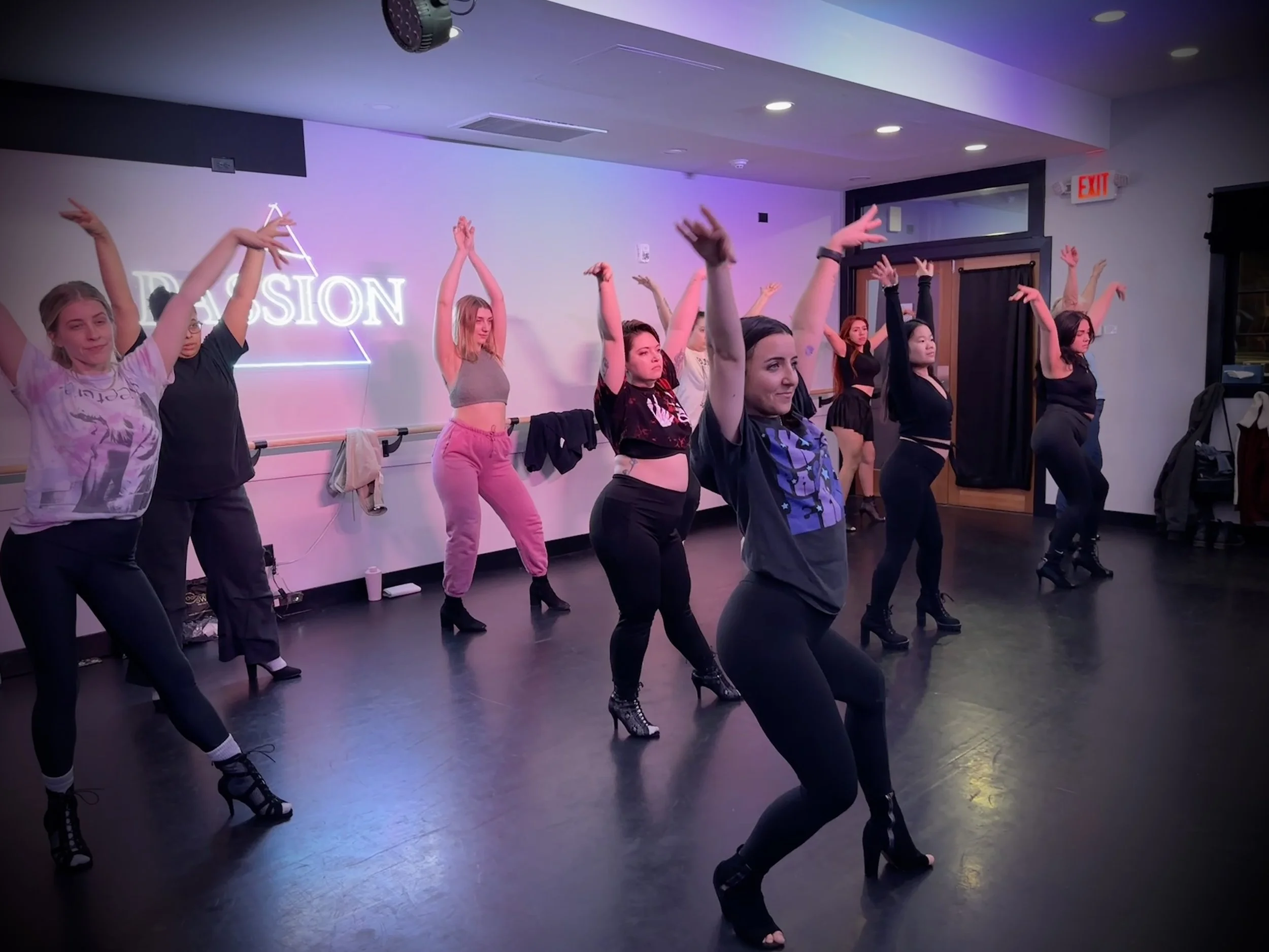 Group of people in black dance attire practicing in a dance studio with wooden floors.