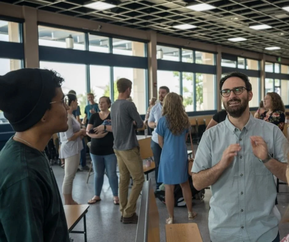 People socializing and conversing in a brightly lit, modern indoor space with large windows, tables, and a grid-pattern ceiling.