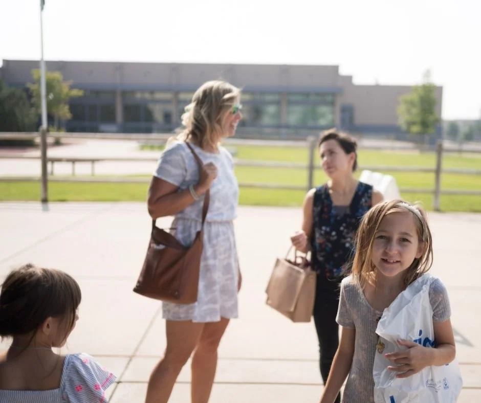 Two women and two young girls stand outside on a sunny day near a fence, with a school building in the background. One woman has curly hair, sunglasses, and a light-colored dress, holding a brown bag. The other woman has dark hair, a dark floral shirt, and is carrying a brown tote. One girl has blonde hair, a striped shirt, and is holding a plastic bag, smiling at the camera. The other girl has dark hair, wearing a light-colored dress with pink and purple flowers, looking away.