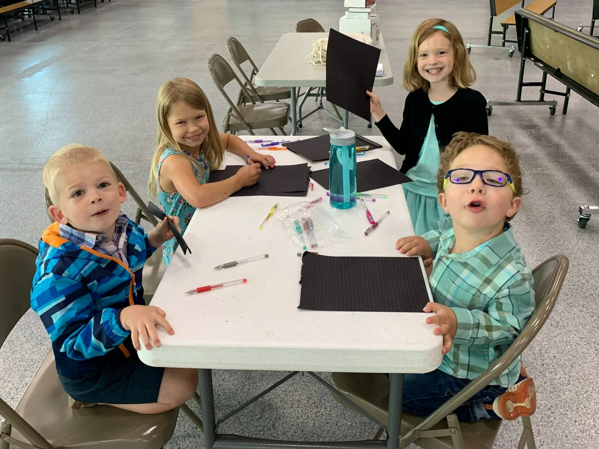 Five young children sitting around a table with black paper, pens, and markers, smiling and engaging in arts and crafts in a classroom or community center.