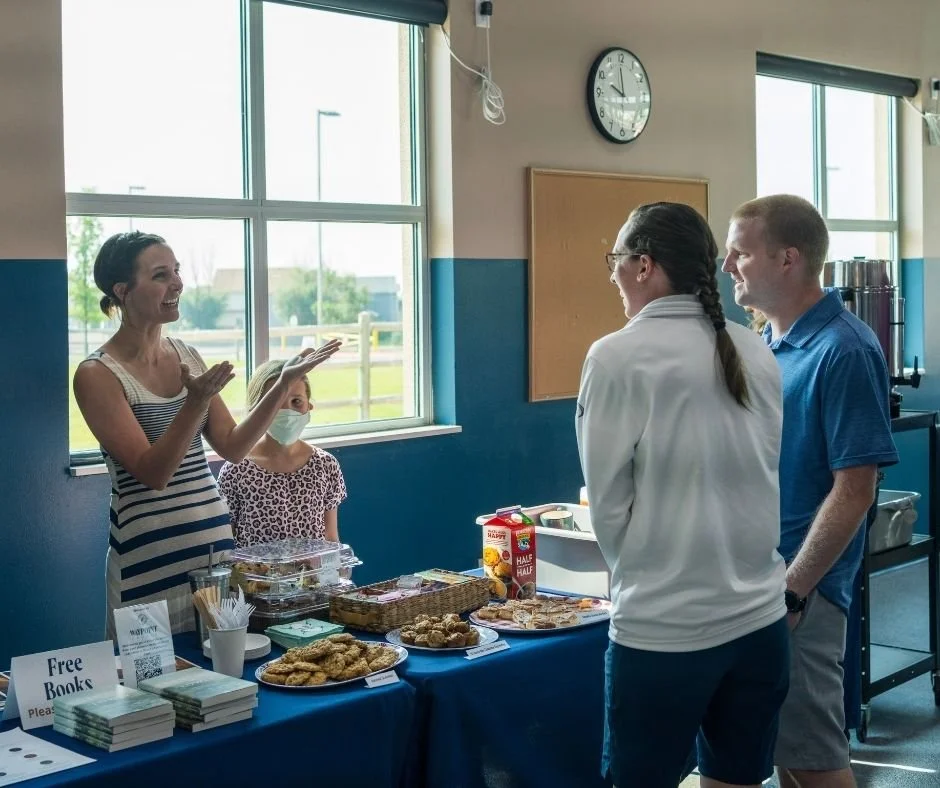 A woman in a striped dress is talking to two people, a woman in a white jacket and a man in a blue shirt, standing in front of a table with food and drinks, inside a room with large windows.