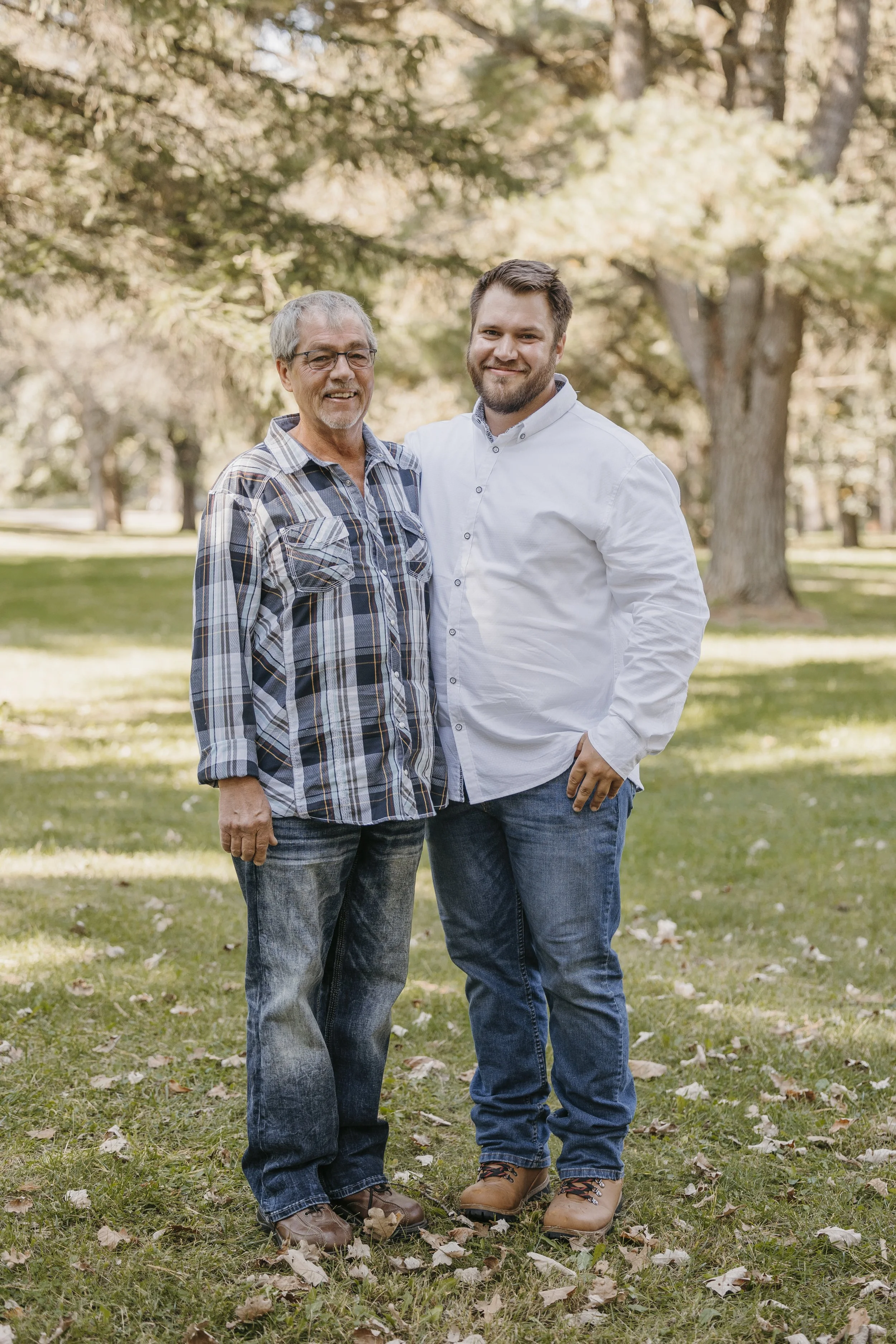 Two men standing in a park, smiling and posing for a photo, with trees and grassy area in the background.