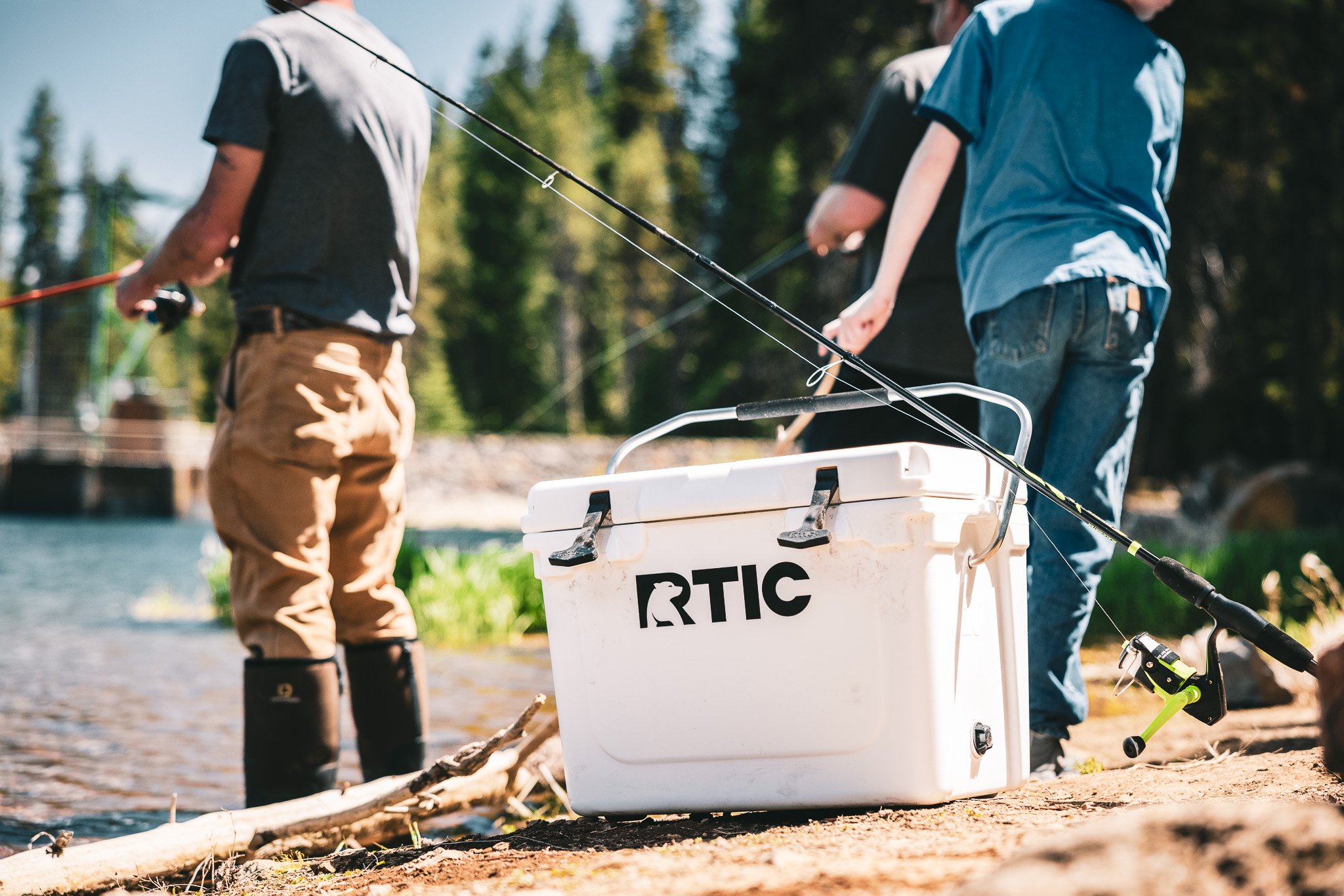 People fishing by a river with fishing rods, a white cooler with the word 'RTIC' and a penguin logo, on a sandy shore with trees in the background.