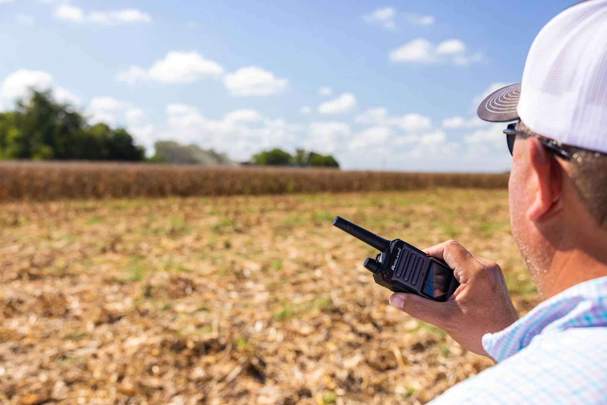 A person wearing sunglasses and a white cap is holding a walkie-talkie or radio device, standing in a field under a blue sky with scattered clouds.