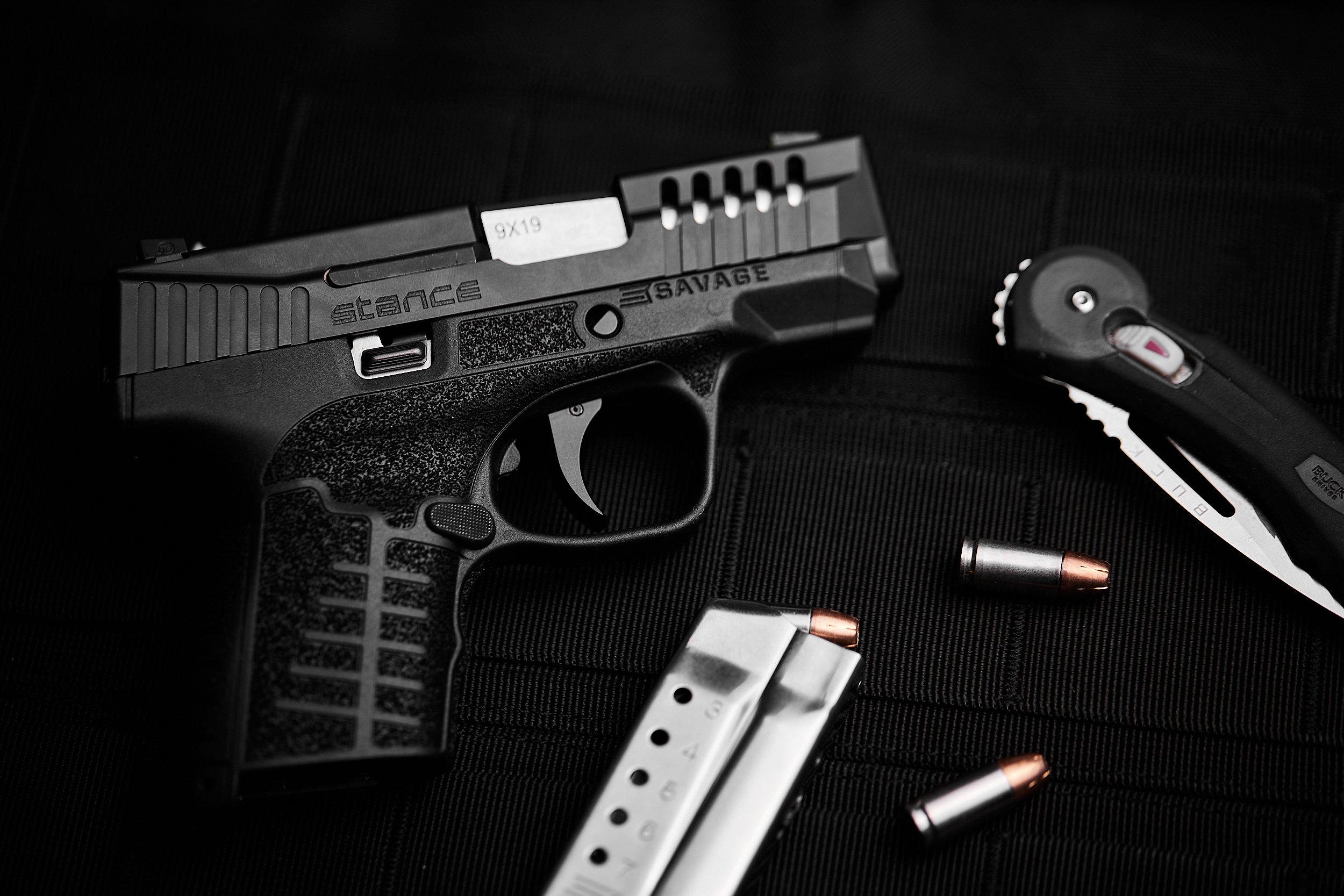 Close-up of a black semi-automatic handgun with spare ammunition and a folding knife on a black surface.
